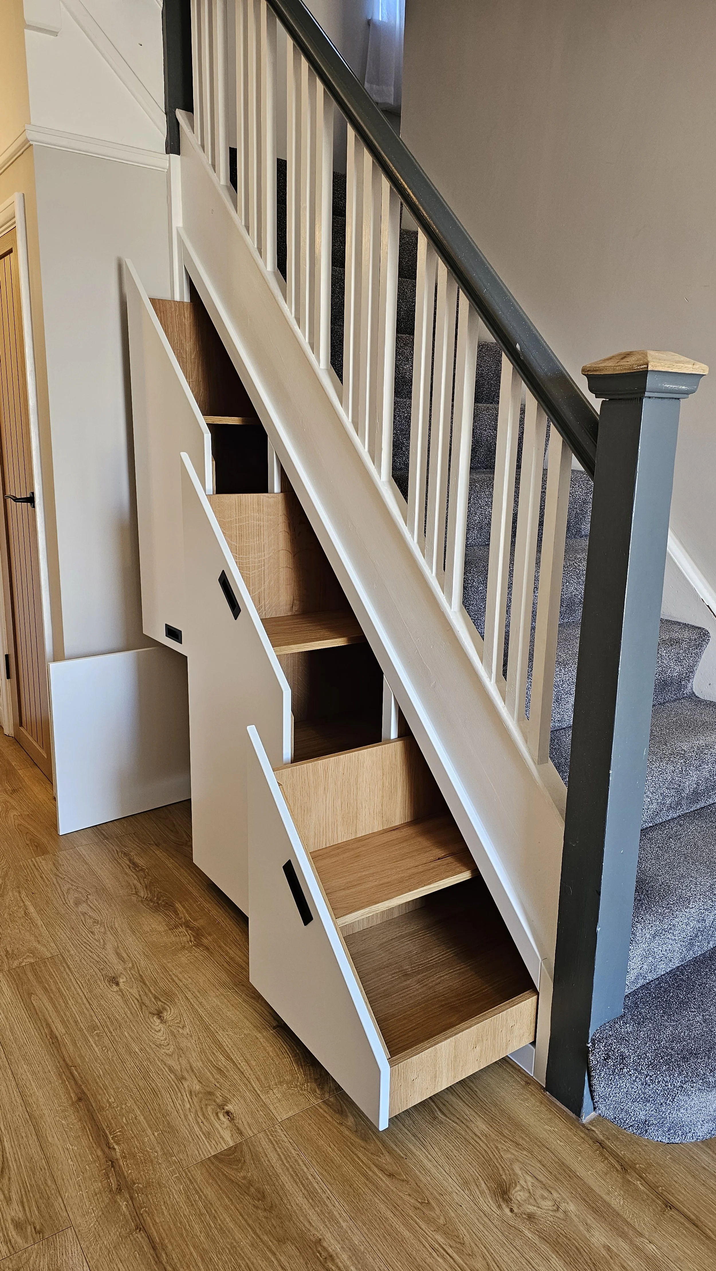 Custom stair storage with shelves built into the lower side of a staircase, featuring black handles, white panels, and wooden shelves.
