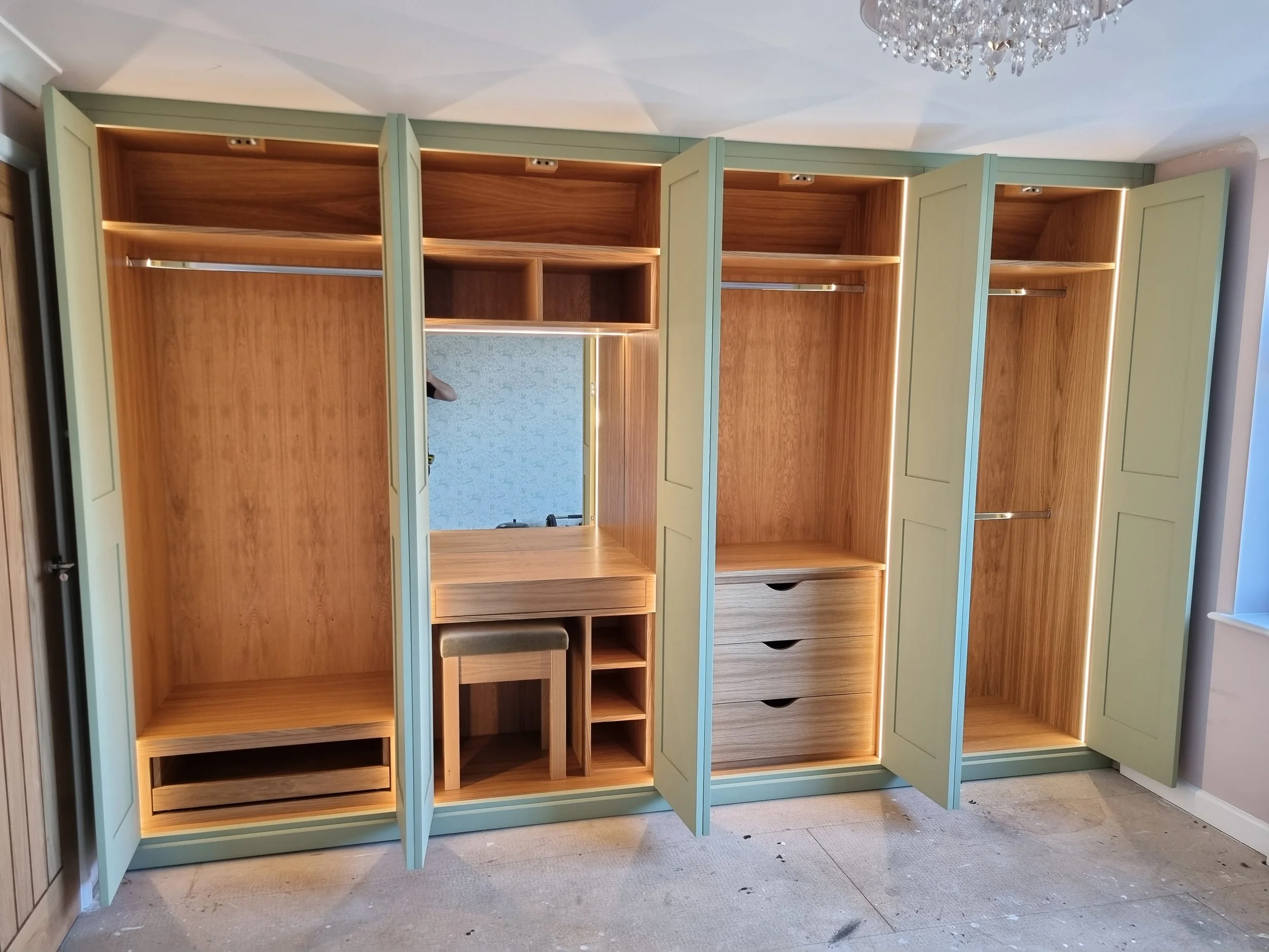 Open green wardrobes with wooden interiors and shelves, drawers, and hanging rods inside, along with a mirrored section and a small desk with a stool.