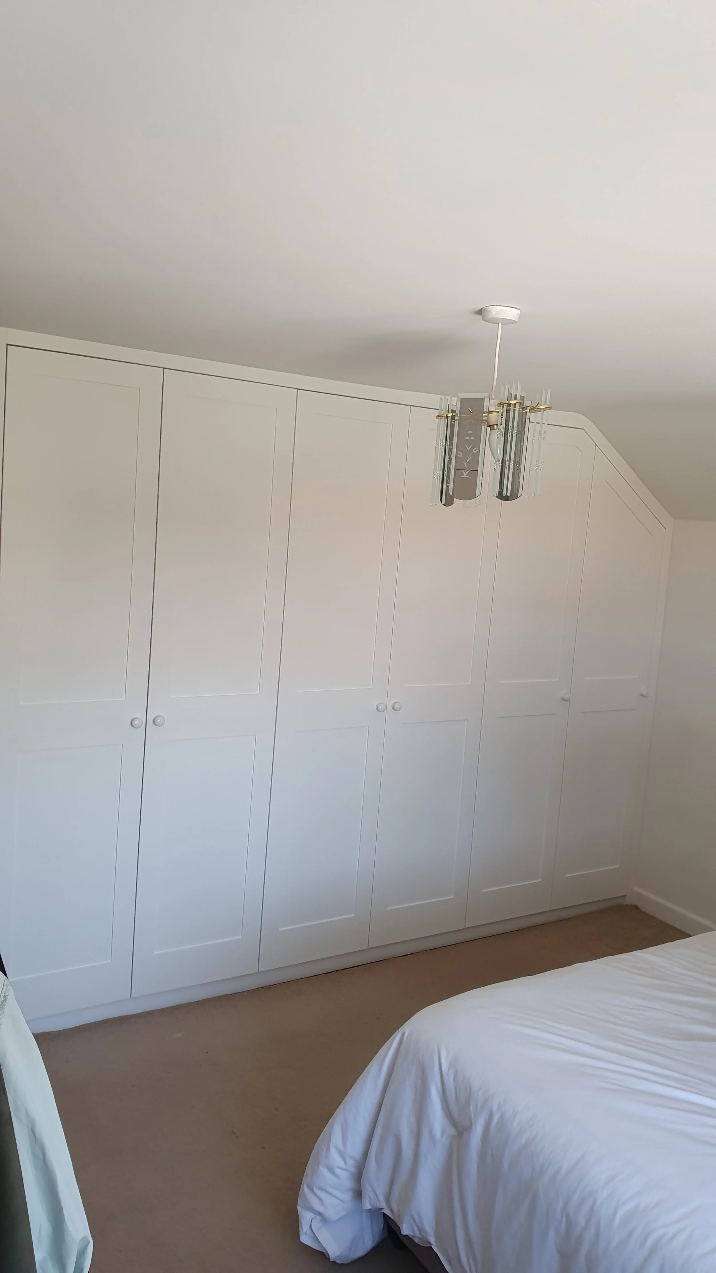 A bedroom with white built-in wardrobes along one wall, a chandelier hanging from the ceiling, and part of a bed with white bedding visible in the foreground.
