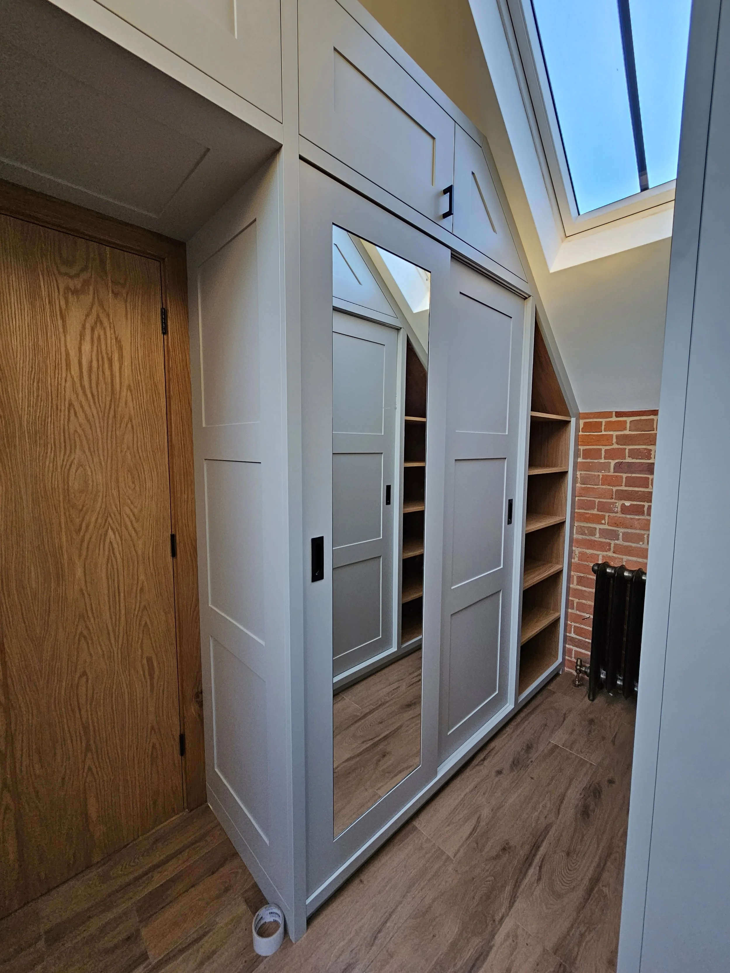 A white wardrobe with sliding doors, one of which has a mirror, situated in a room with wooden floors and a brick wall. There are shelves inside the wardrobe, and a skylight visible above.