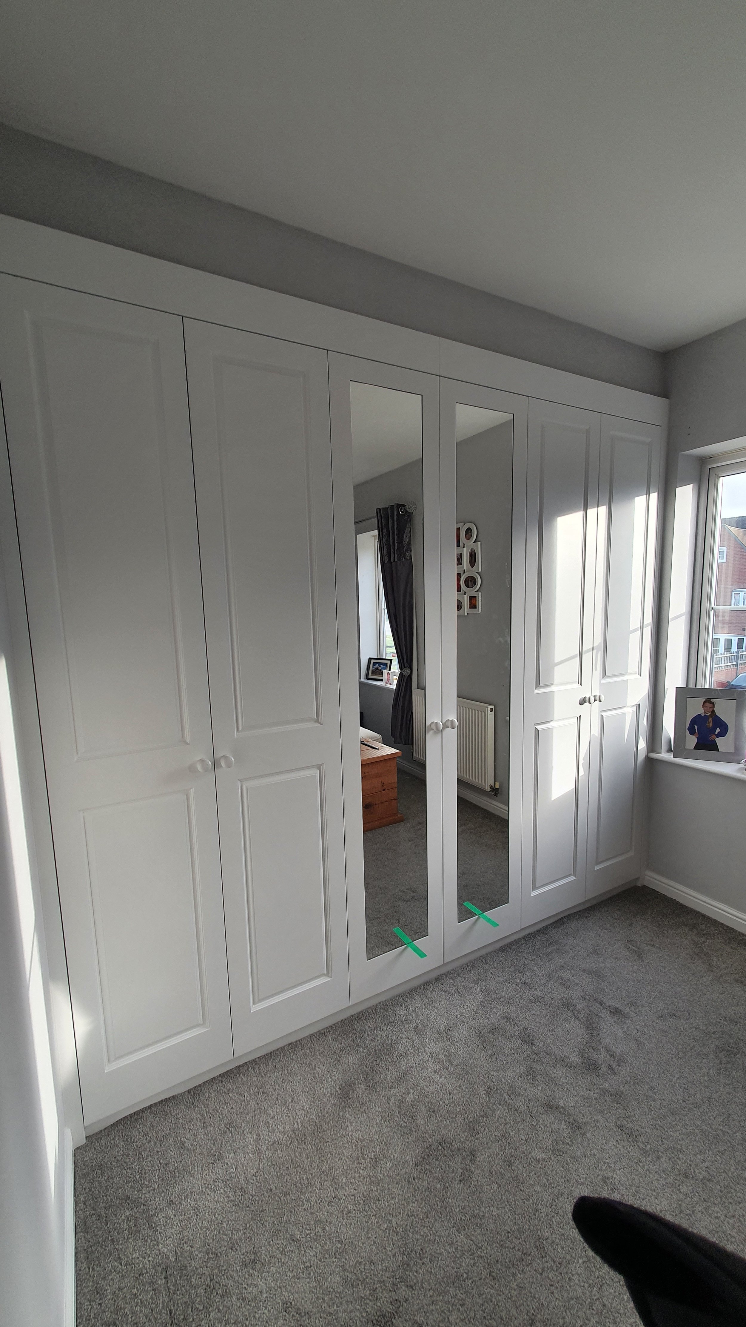 White built-in wardrobe with two mirrored doors in a bedroom, sunlight on gray carpet, window on the right, framed photo on windowsill.