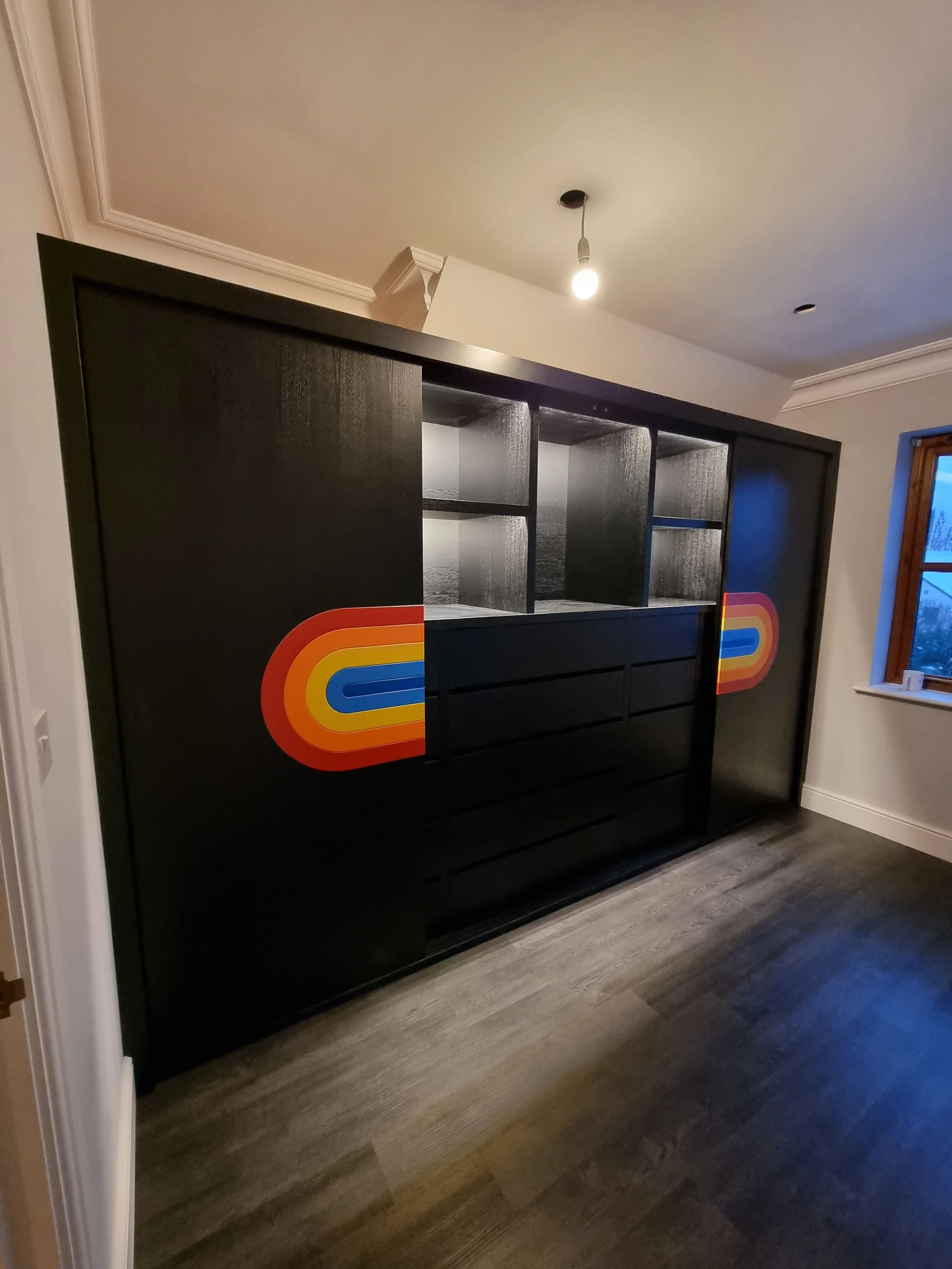 A black sliding door wardrobe with rainbow-colored semi-circular design on either side, situated in a room with wood flooring, a window, and a ceiling light