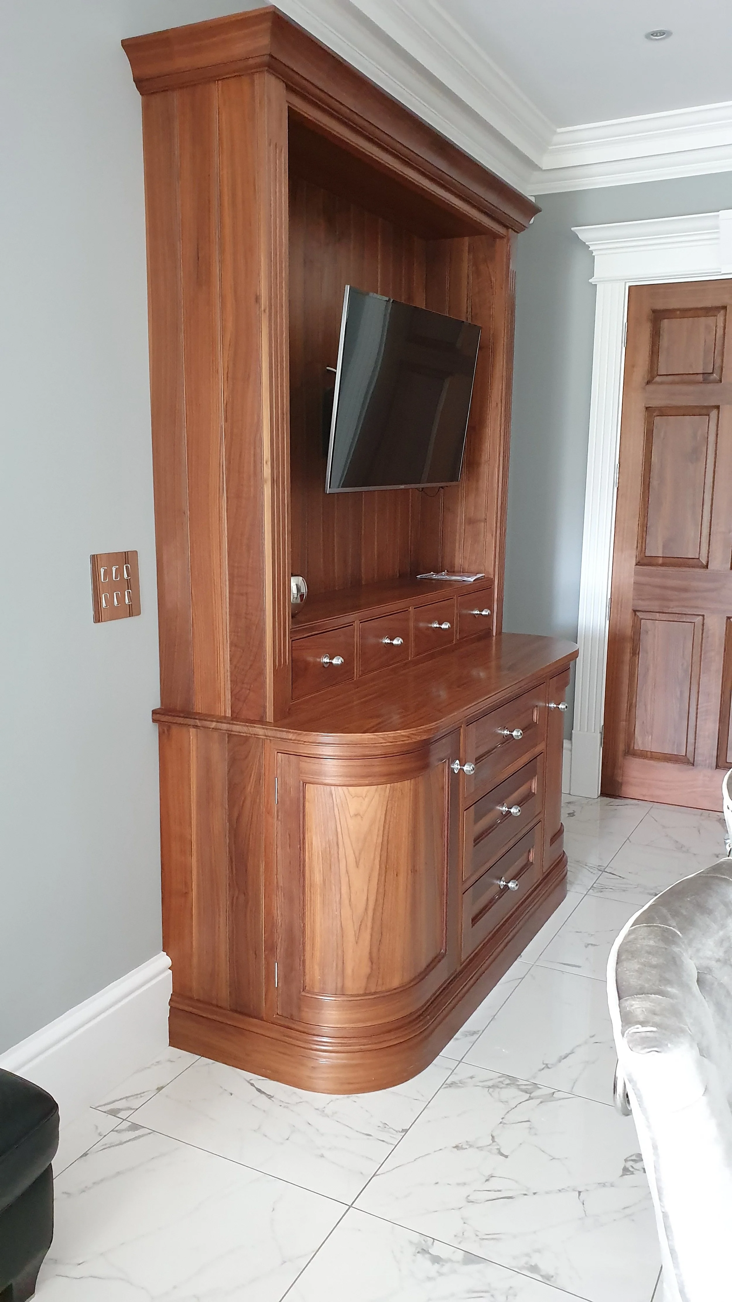 A wooden entertainment center with a mounted flat-screen TV and multiple drawers, located in a room with marble tile floor and white crown molding.