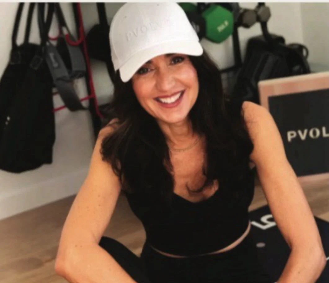 A woman with long dark hair wearing a white cap and black sleeveless top, smiling and sitting on the floor in a gym or fitness area.