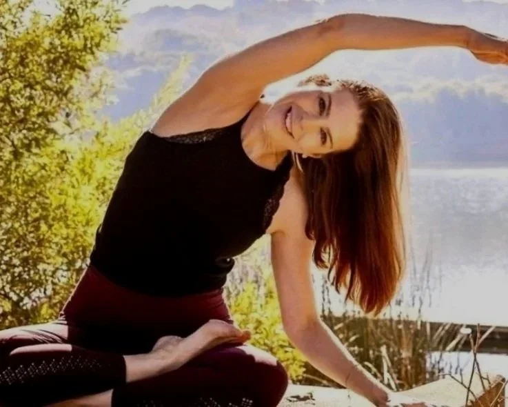 Woman practicing yoga outdoors near a lake with trees in the background, performing a side stretch pose.