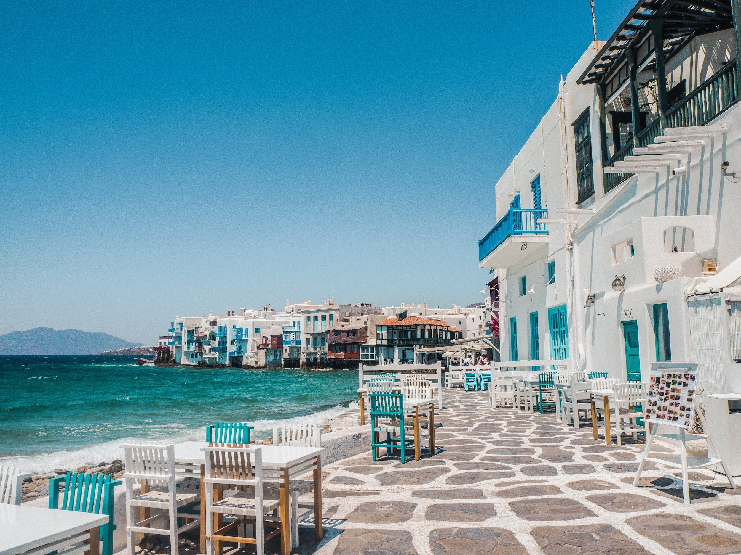 Seaside view of Mykonos with white and blue buildings, tables and chairs on a stone-paved walkway, and the Aegean Sea.