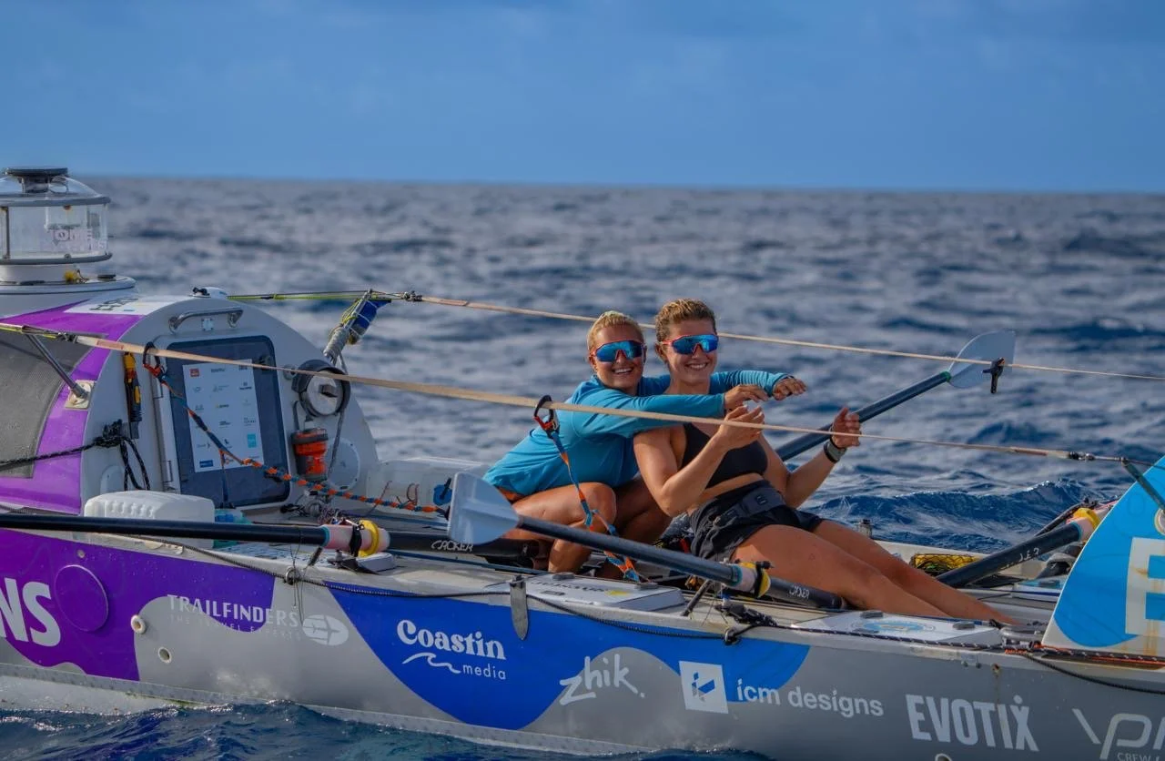 Two women (Jess and Mims from Seas The Day, sailing in a small boat on the Pacific ocean, wearing sunglasses and life jackets, smiling and holding paddles.