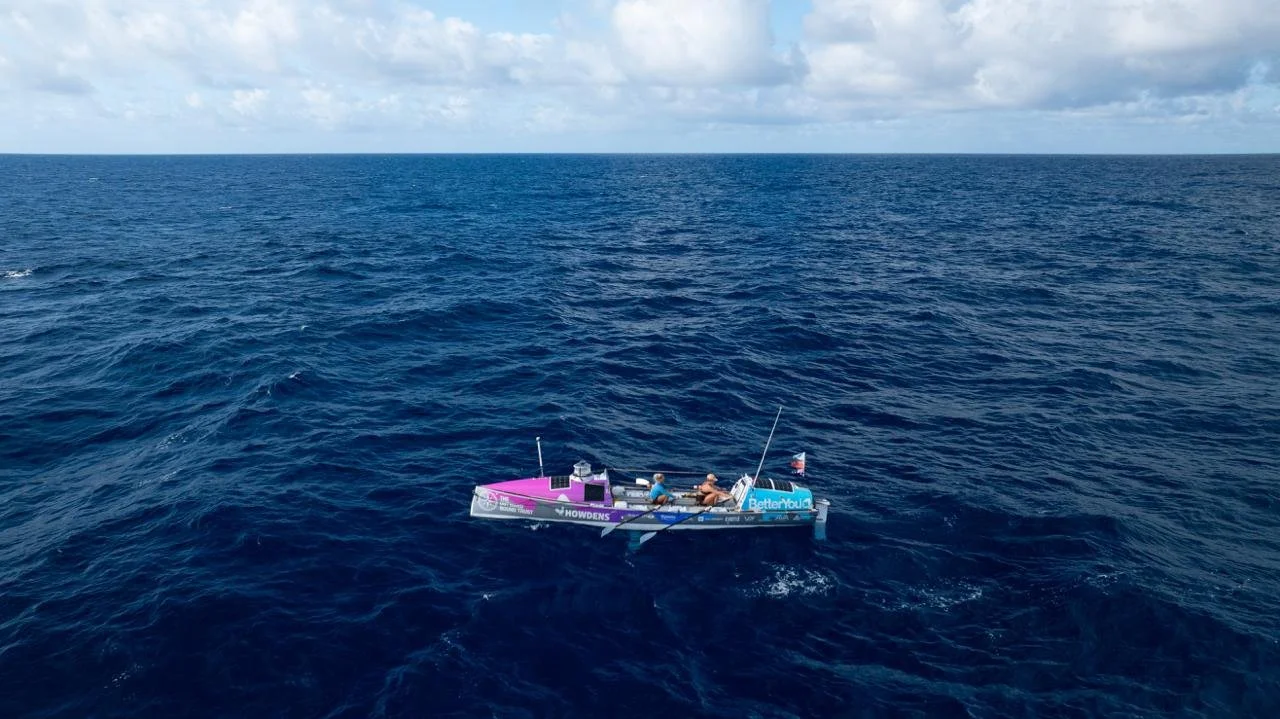 Two people in a small race boat on the open ocean, with a clear sky and some clouds above.