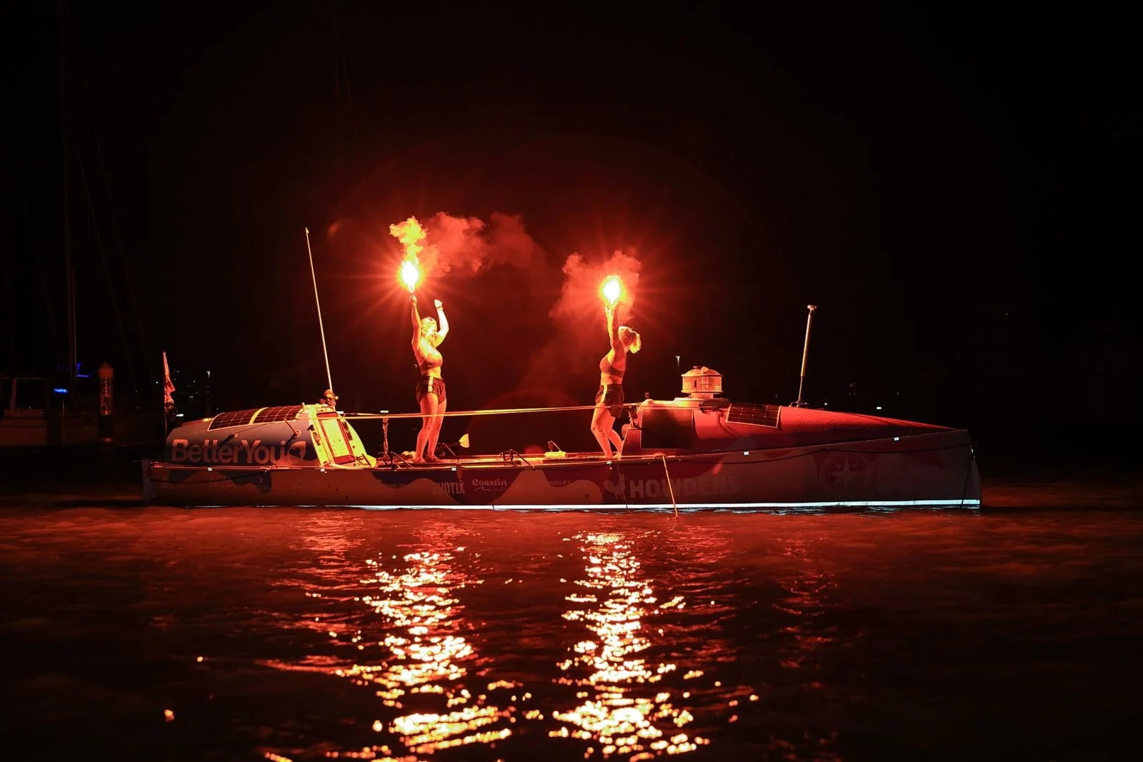 Two women on a boat at night holding flares that emit bright red light, reflecting on the water.