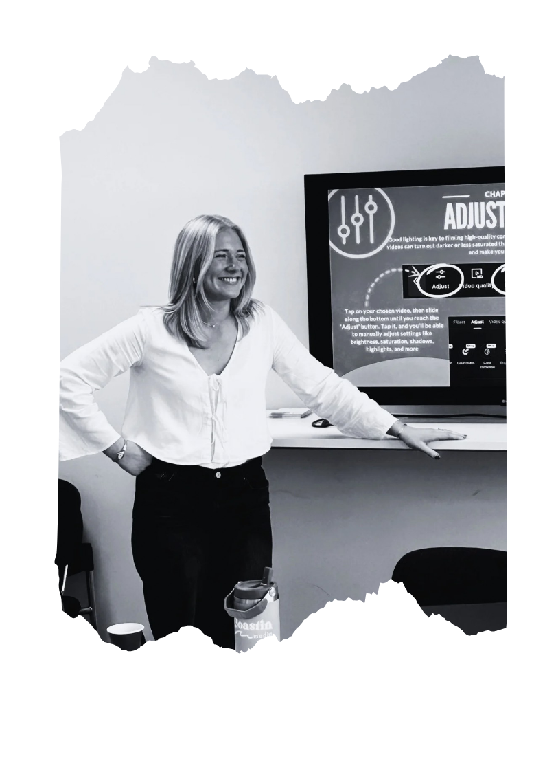 Jodie standing in front of a screen displaying video editing instructions, smiling with one hand on the table and the other on her hip, in a classroom or conference room setting.