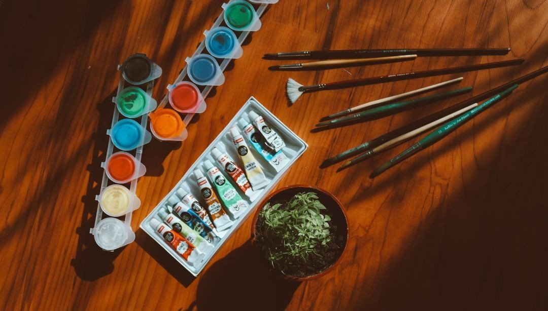 An image of paint sets with brushes on a wooden table next to a small plant.