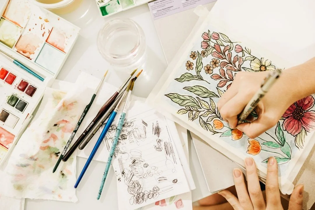 An image of someone drawing flowers and leaves in a sketchbook with a pen surrounded by brushes, a cup of water, and a paint pallet.