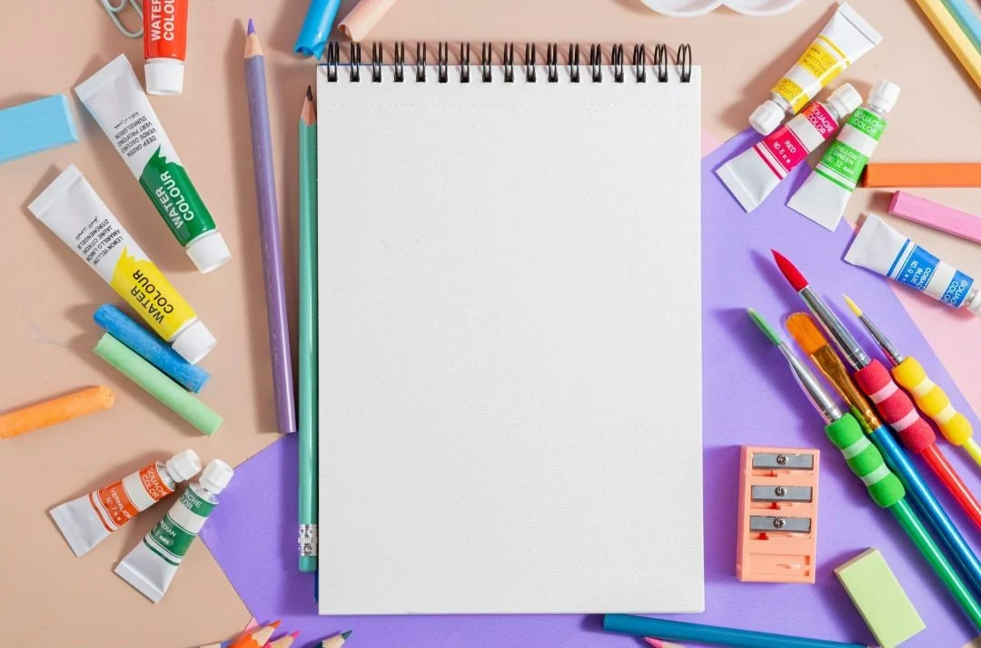 A sketch pad surrounded by art materials, including paints, colored pencils, brushes, and a pencil sharpener.