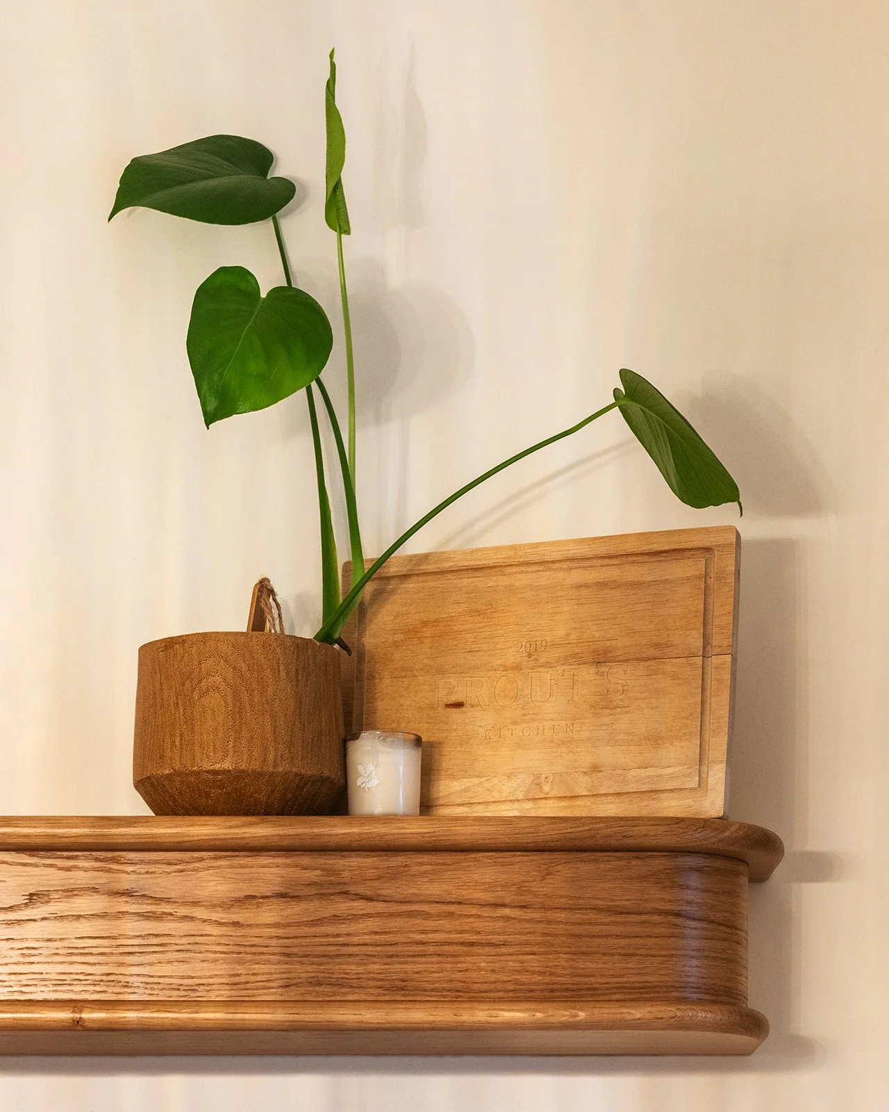 A wooden shelf holds a potted plant with large green leaves, a small white candle, and a wooden cutting board with the engraved words '2019 PROUTE'S KITCHEN' against a plain light-colored wall.