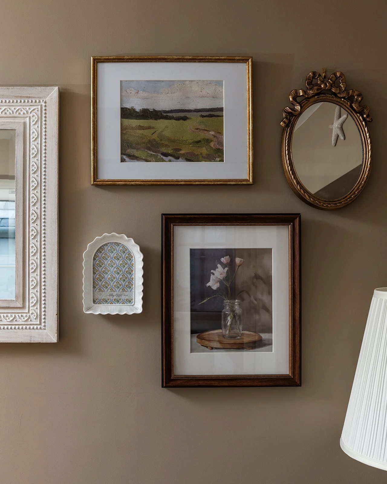 A gallery wall with five framed pictures and a mirror, including landscape paintings, floral photograph, and decorative art, in a cozy living room corner.