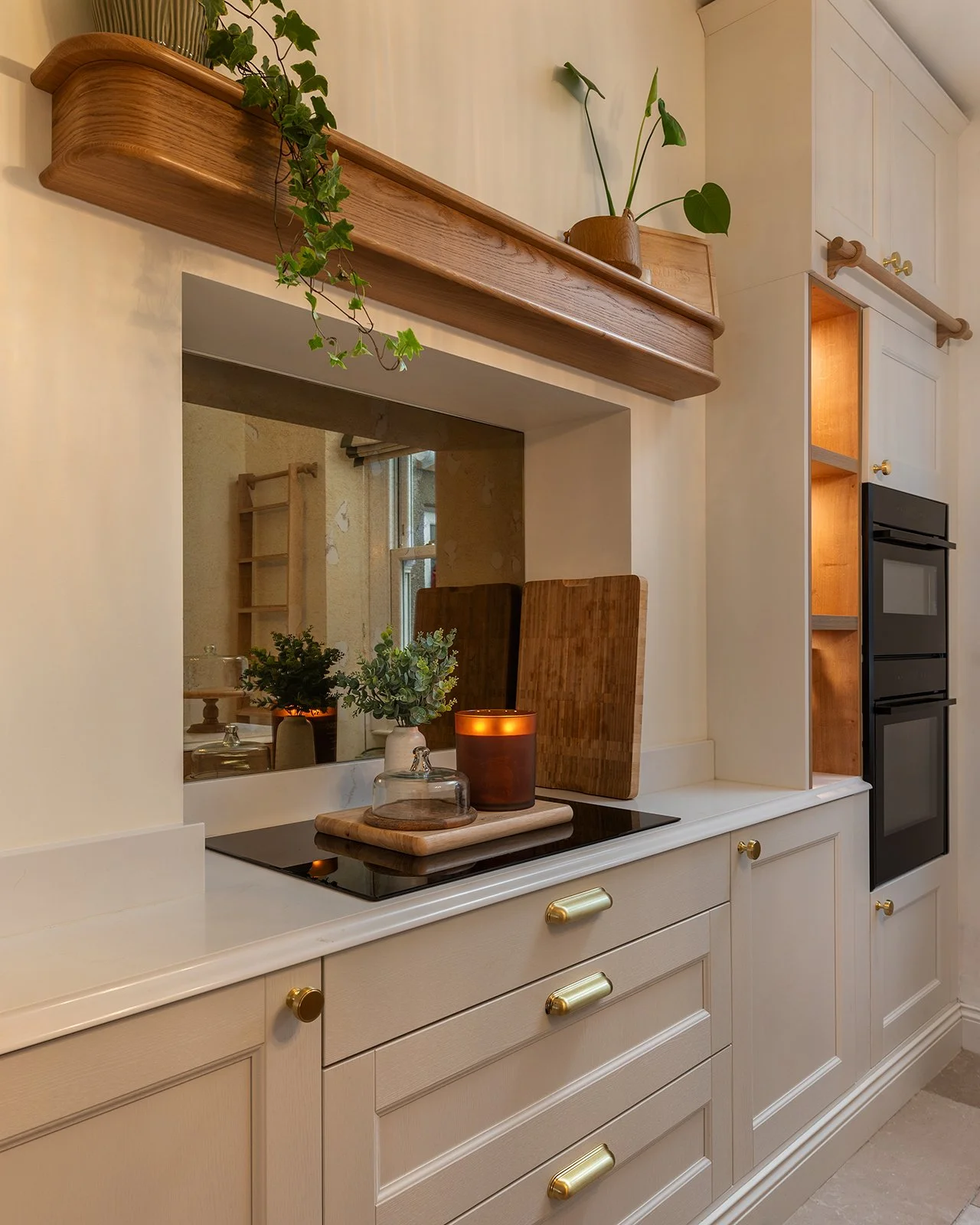 A kitchen counter with a black electric stove, a small glass cloche, a cutting board, a candle, and green plants in small vases. There is a large mirror behind the counter and a wooden shelf above with a potted plant.
