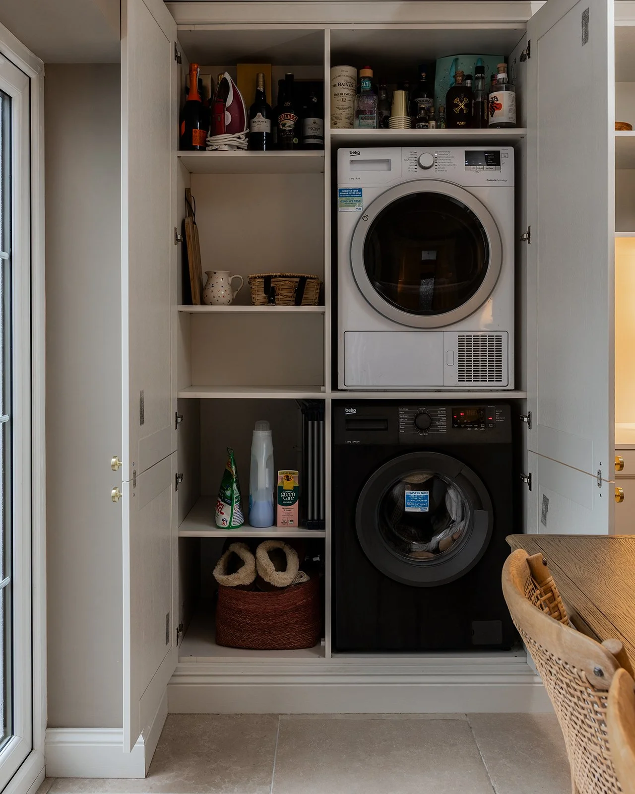 Laundromat laundry closet with a stacked washer and dryer inside a cabinet with open doors, shelves with bottles and kitchen items, and a basket with slippers on the bottom shelf.