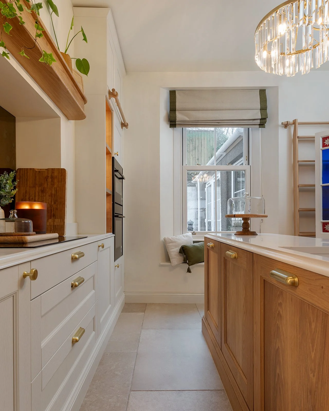 Kitchen with white and wooden cabinets, a window with a green and beige roman shade, a wooden candle holder with a glass dome, and a chandelier overhead.