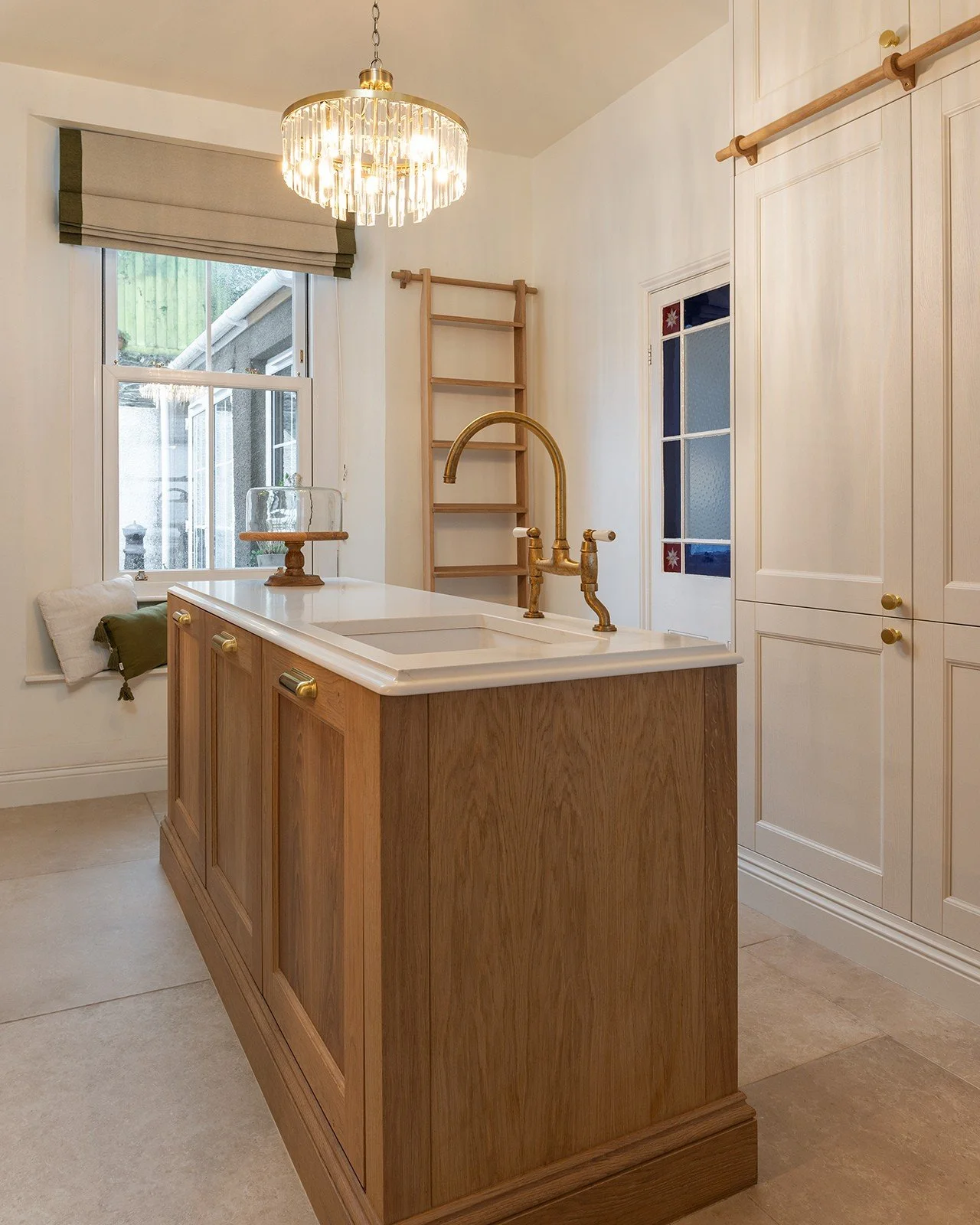 Kitchen island with wooden cabinetry, white countertop, and brass faucet, near a window with a beige Roman shade, a chandelier overhead, and white cabinets with brass knobs on the right side.