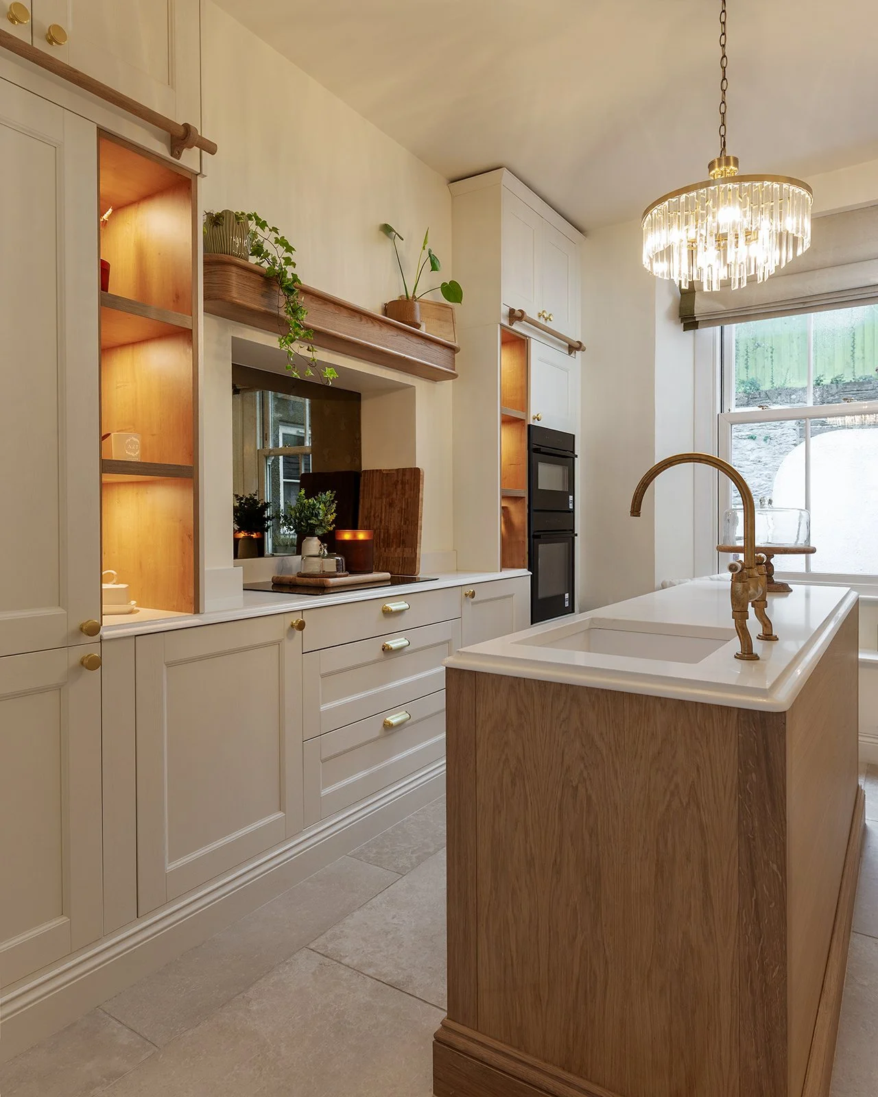 A modern kitchen with white cabinets, a wooden kitchen island with a sink, a chandelier, and a large window with blinds.