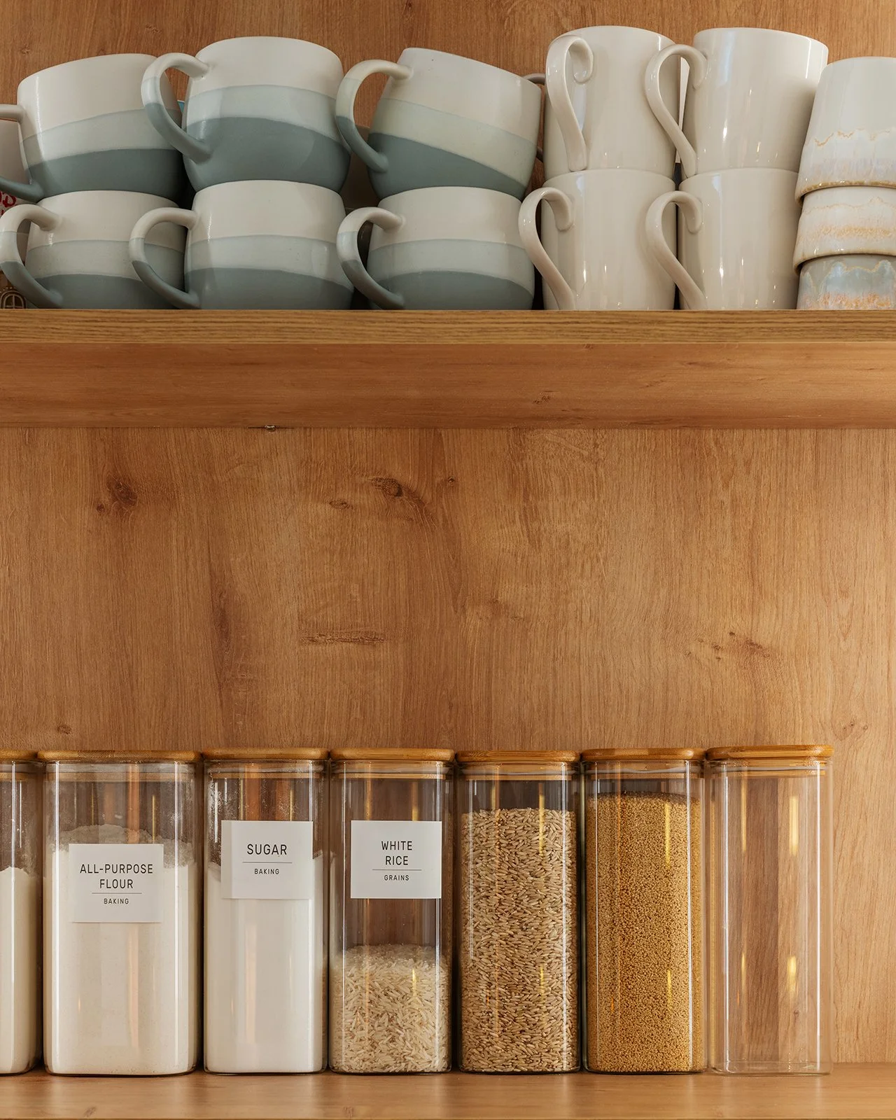 White ceramic coffee mugs with gray and cream accents on a wooden shelf above clear containers of baking ingredients like flour, sugar, rice, and grains.
