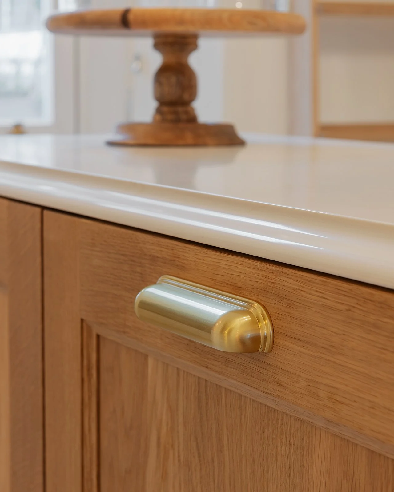 Close-up of a kitchen cabinet with a wooden finish and a gold-colored handle, with a cream-colored countertop and a wooden table in the background.