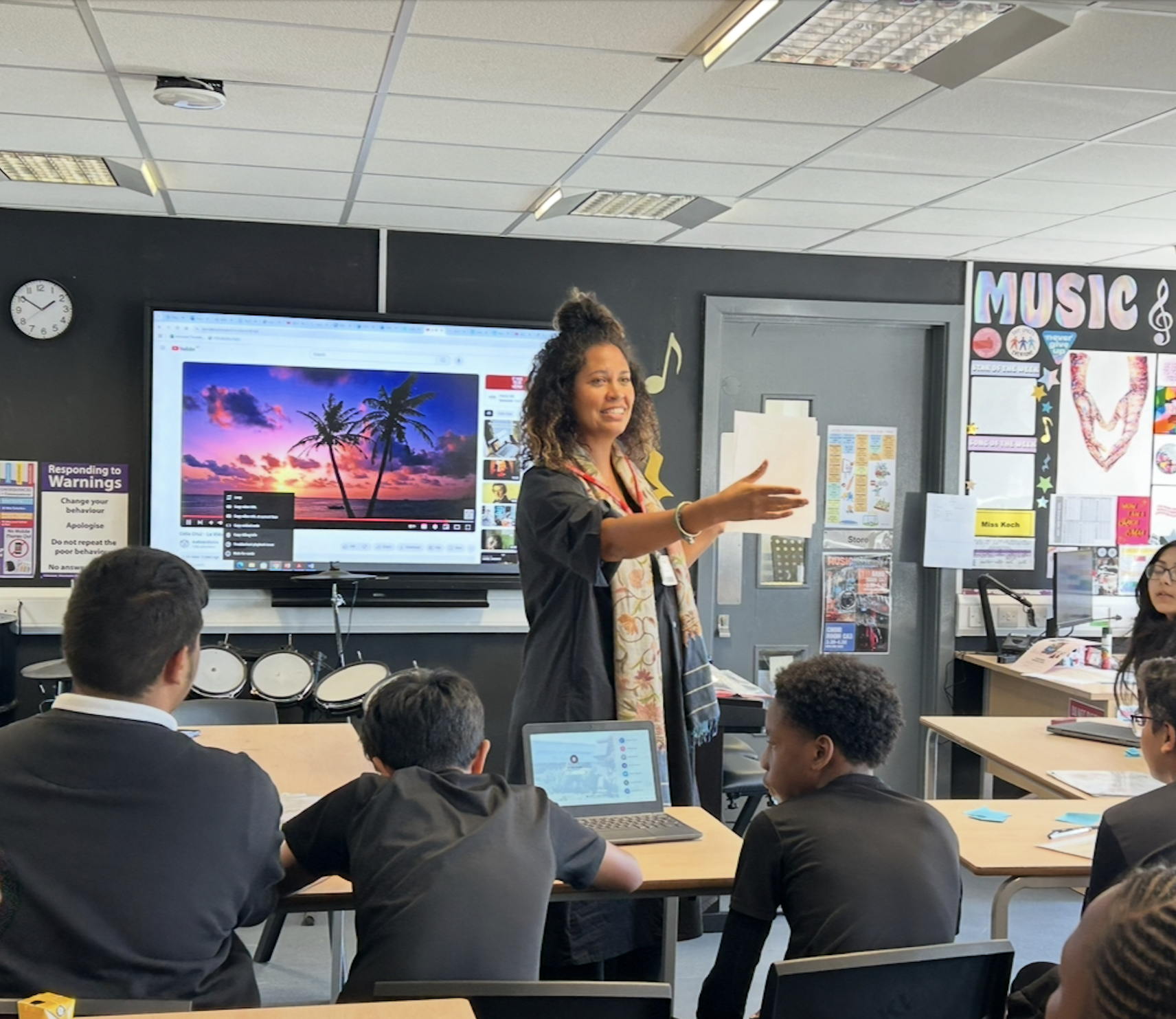 A teacher standing in front of a classroom with students, pointing at a large screen displaying a sunset over palm trees. The classroom is decorated with musical notes and music-themed posters.