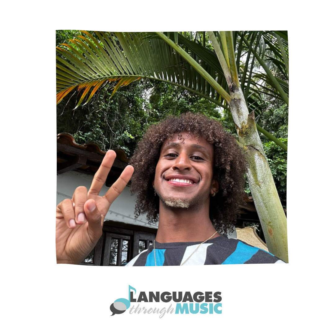 A person with curly hair smiling and making a peace sign in front of a tropical background with palm trees.