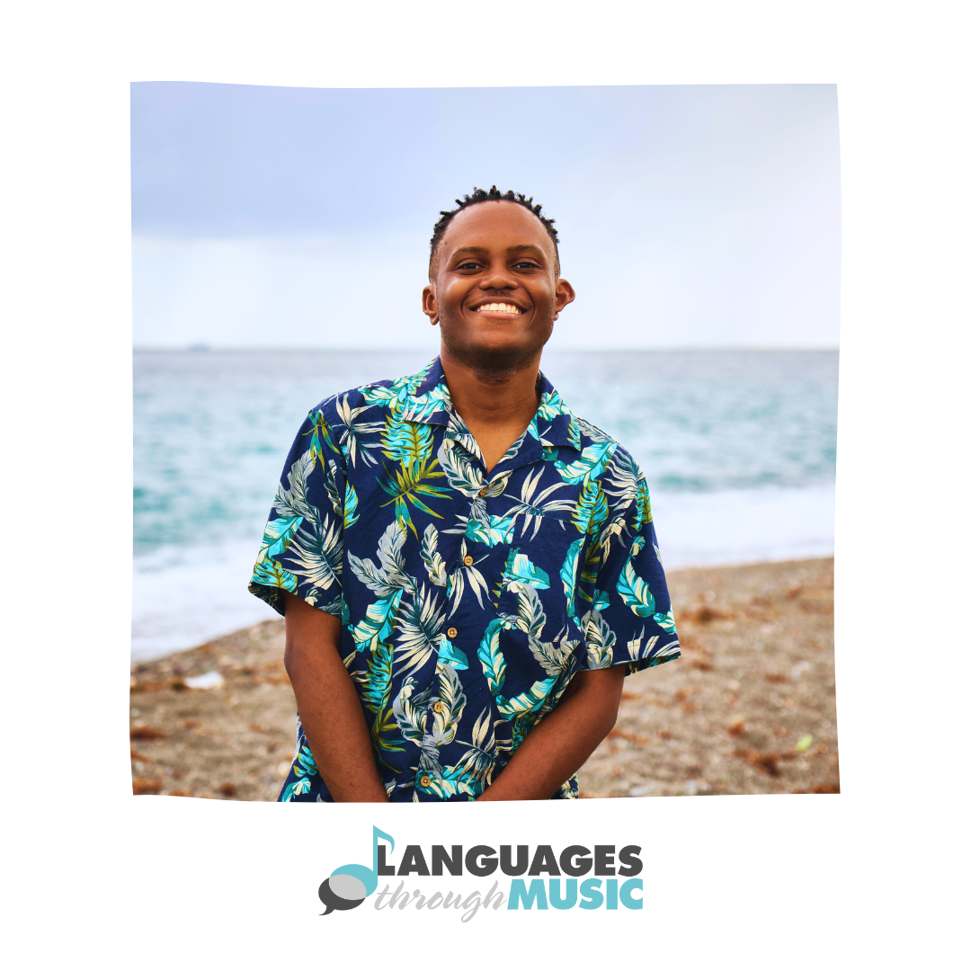 A smiling man in a blue Hawaiian shirt standing on a beach near the ocean with a cloudy sky in the background.