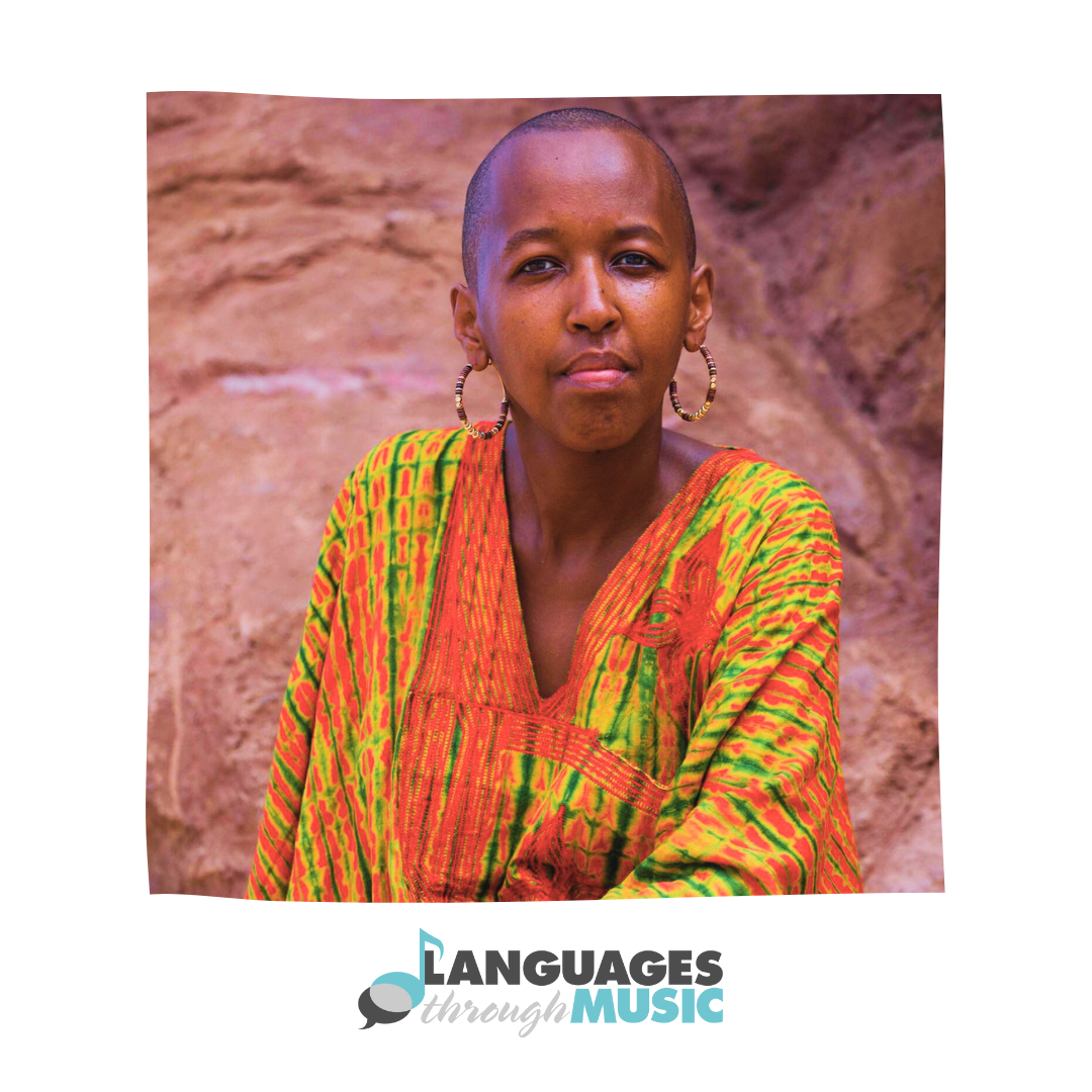 A woman with a shaved head wearing large hoop earrings and a colorful, patterned traditional dress against a natural, rocky background.