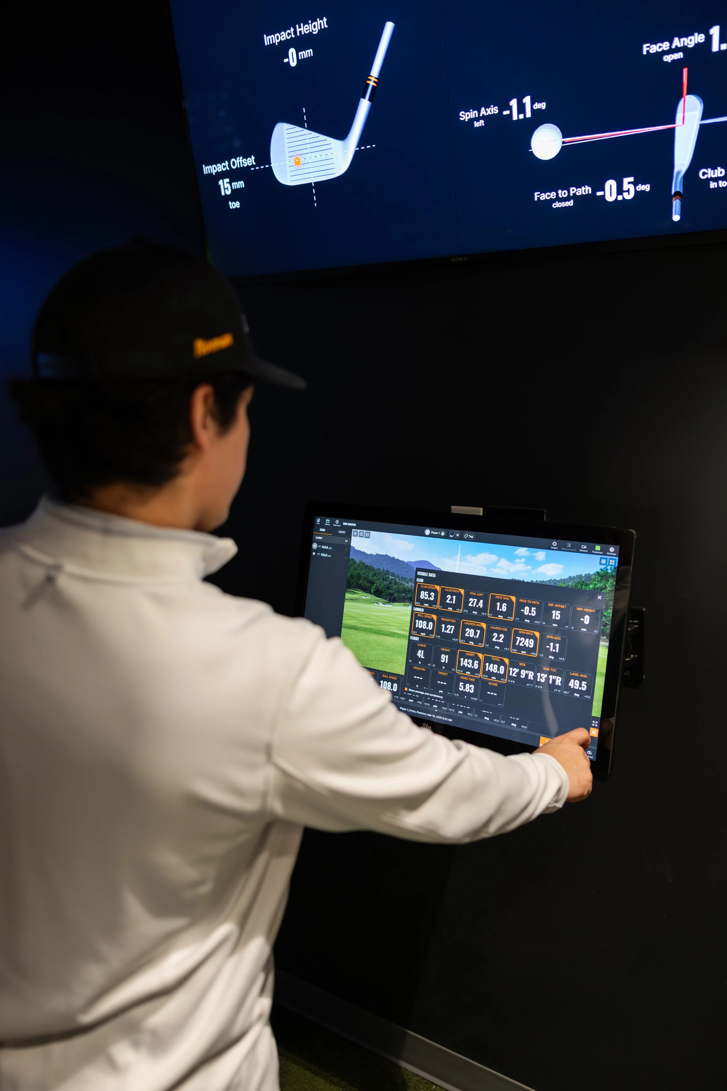 A person operating a golf simulation system with a monitor showing golf swing data and a virtual golf course background.