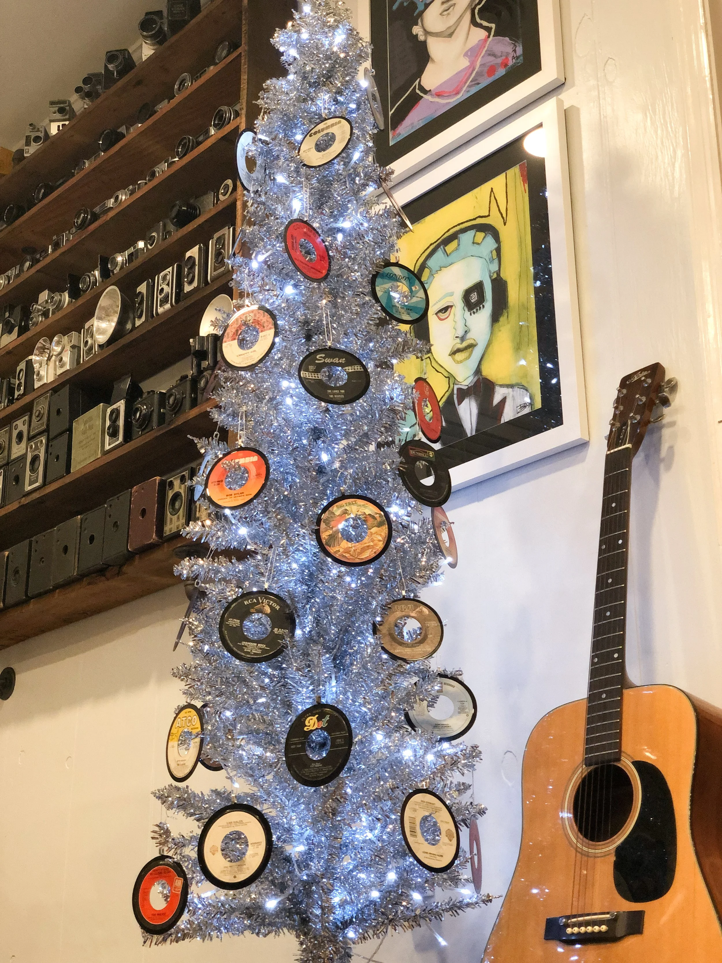 A silver tinsel Christmas tree decorated with vinyl records, set against a wall with framed art and a mounted collection of vintage cameras. A guitar leans against the wall nearby.