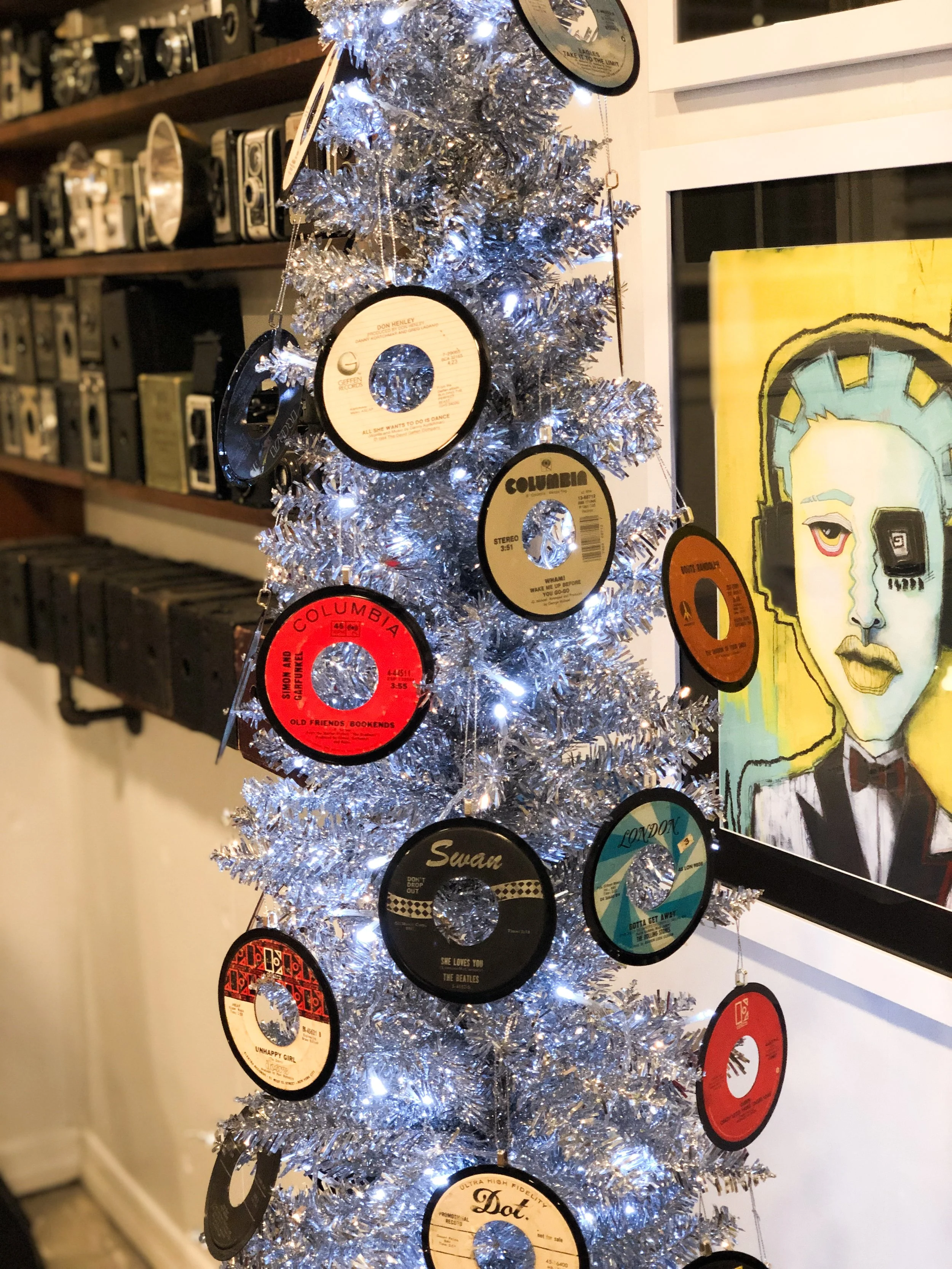 Silver Christmas tree decorated with vintage vinyl records and lights, surrounded by old cameras and a modern art painting in the background.