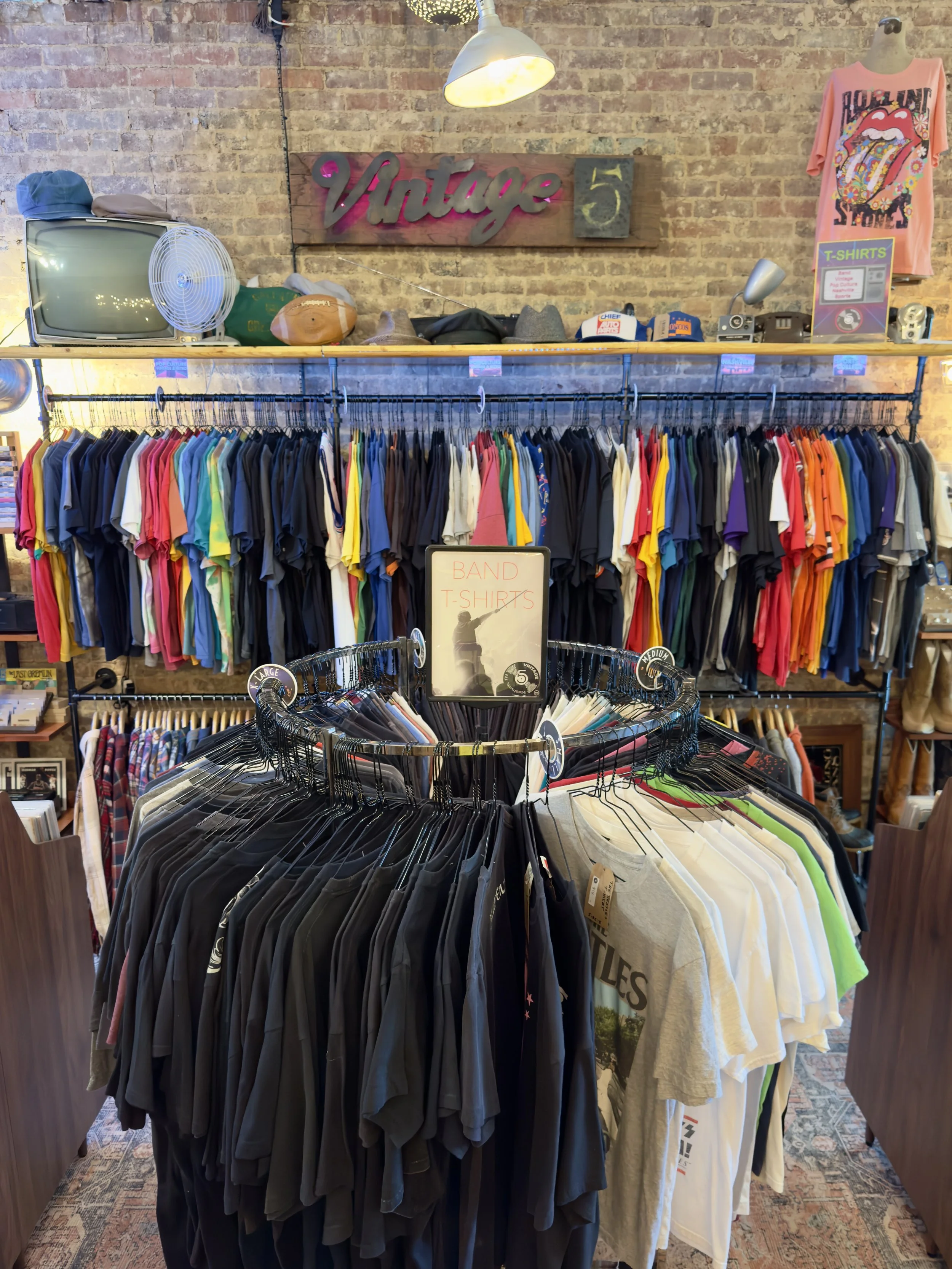 Clothing store display with racks of Band T-Shirts, Vintage T-shirts in various colors, a sign reading 'Vintage 5,' a vintage television, hats, a fan, and a wall with exposed brick and a neon sign.