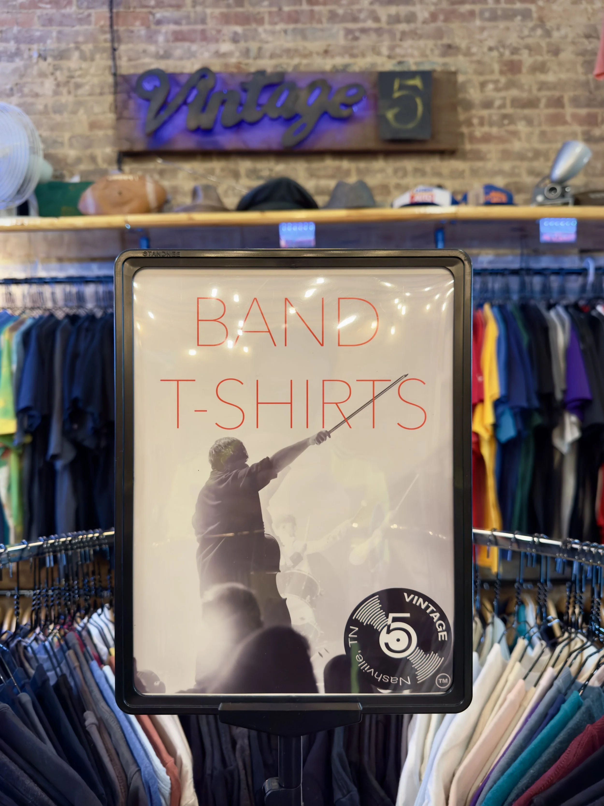A clothing store display with a sign advertising band T-shirts. The sign features an image of a man playing a guitar and has a circular sticker indicating vintage clothing.