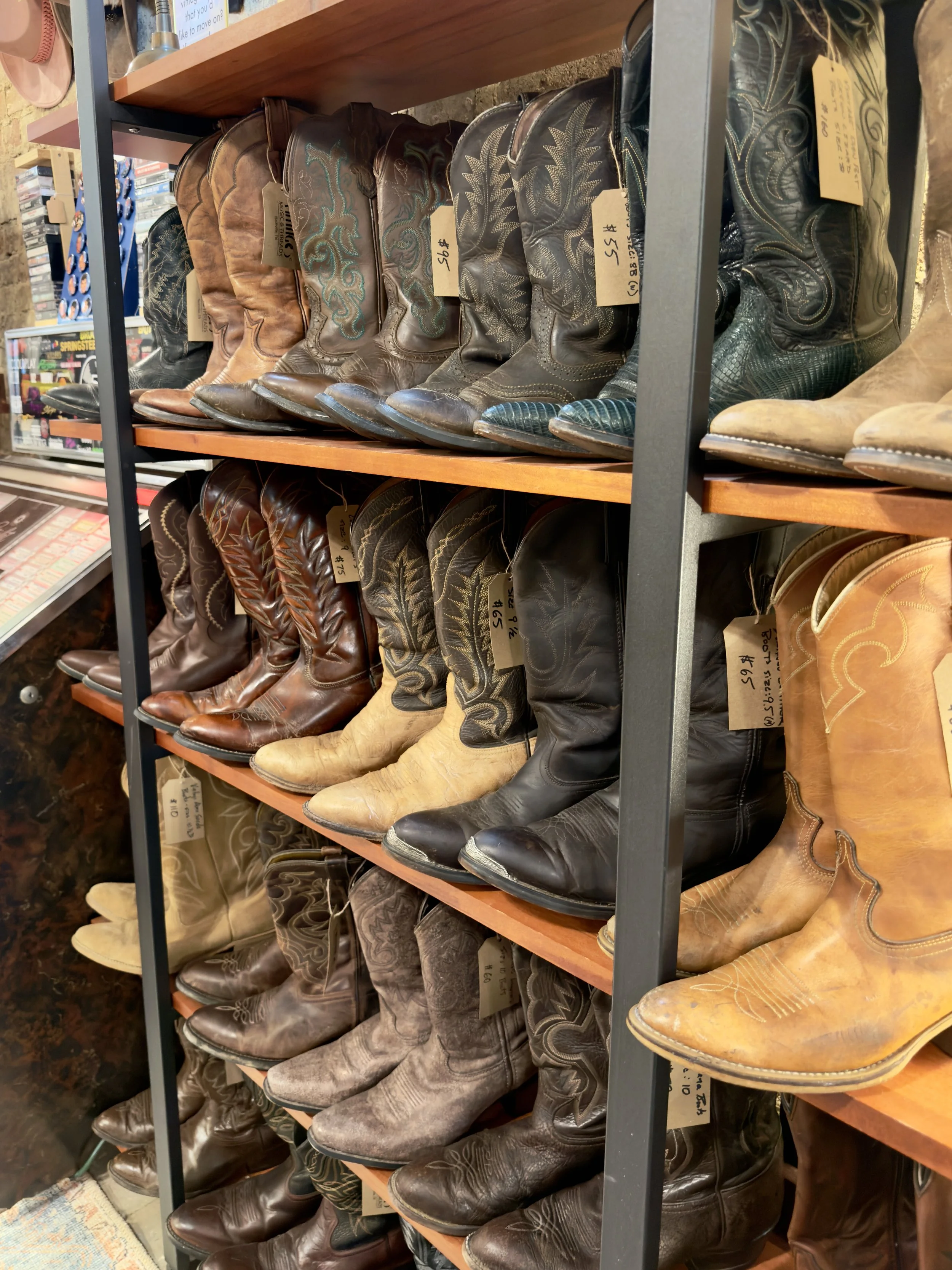 A variety of men's and women's, vintage and new, leather cowboy boots on wooden shelves in a store.