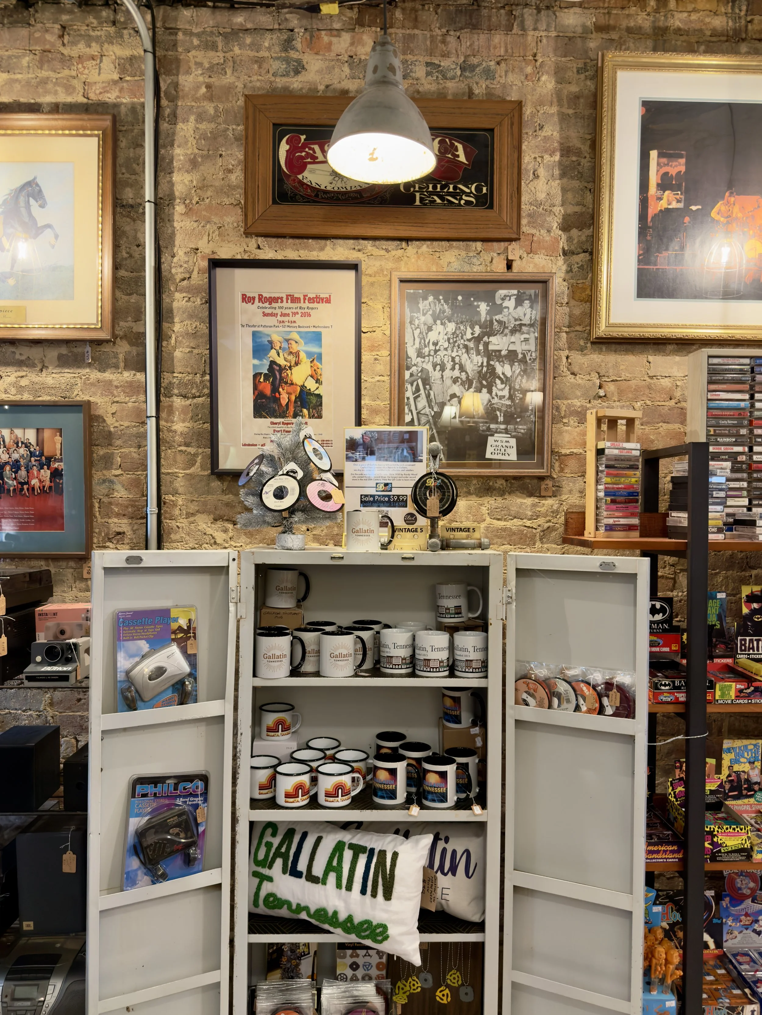 A brick wall inside a store with framed movie, concert, Nashville and western posters and photographs, and shelves filled with various Gallatin, TN merchandise including mugs, figurines, and accessories.