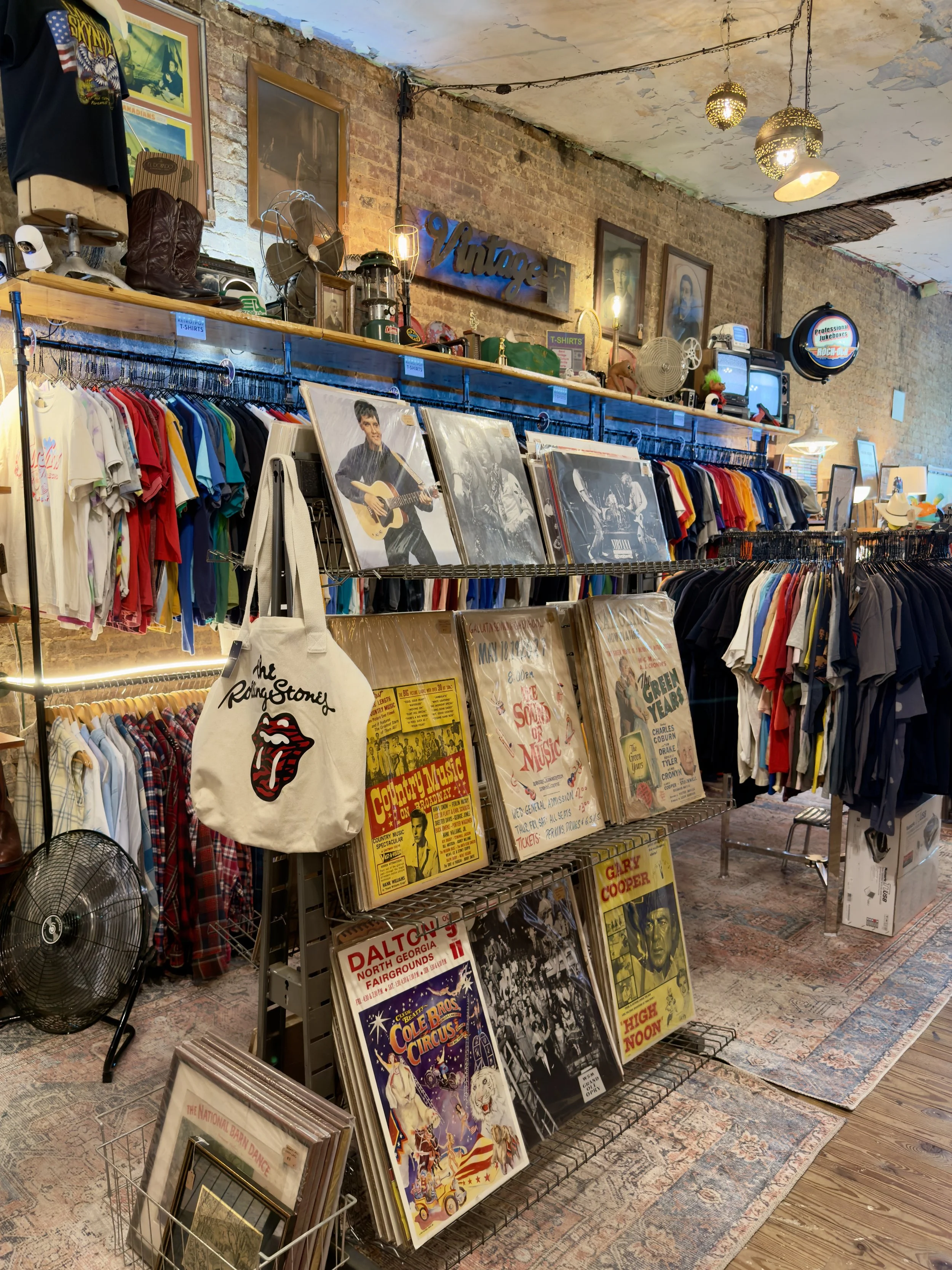 Inside a vintage store with clothing racks with vintage clothes and band t-shirts. Various memorabilia and framed movie and concert posters are arranged on shelves and the wall, along with other vintage items.