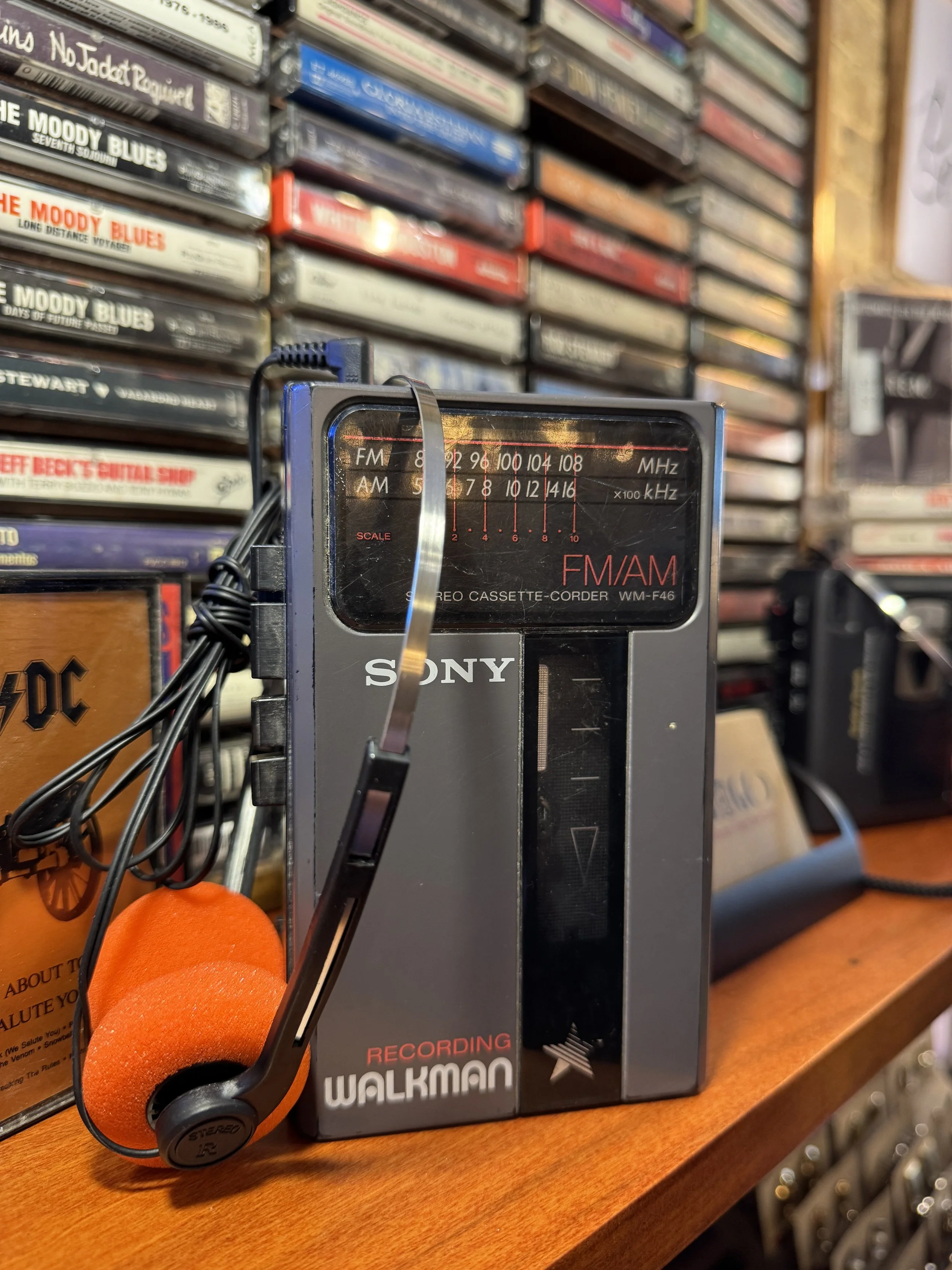 Close-up of a vintage Sony Walkman portable cassette player with a pair of black and orange headphones resting on a wooden surface. In the background, shelves filled with cassette tapes are visible, creating a nostalgic music scene.