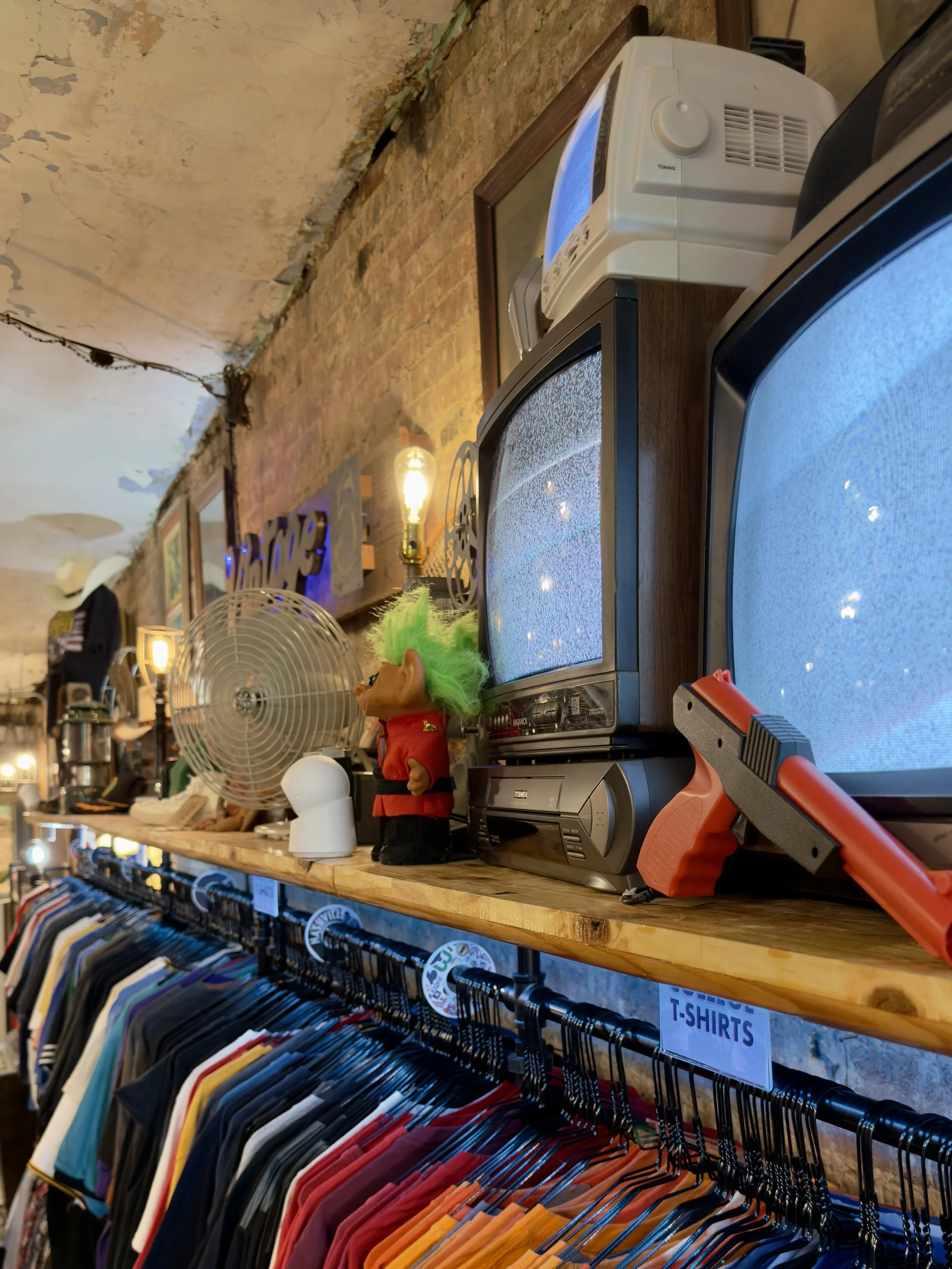 Clothing rack displaying vintage and band t-shirts in a store with vintage decor, fans, and vintage televisions on a wooden shelf.