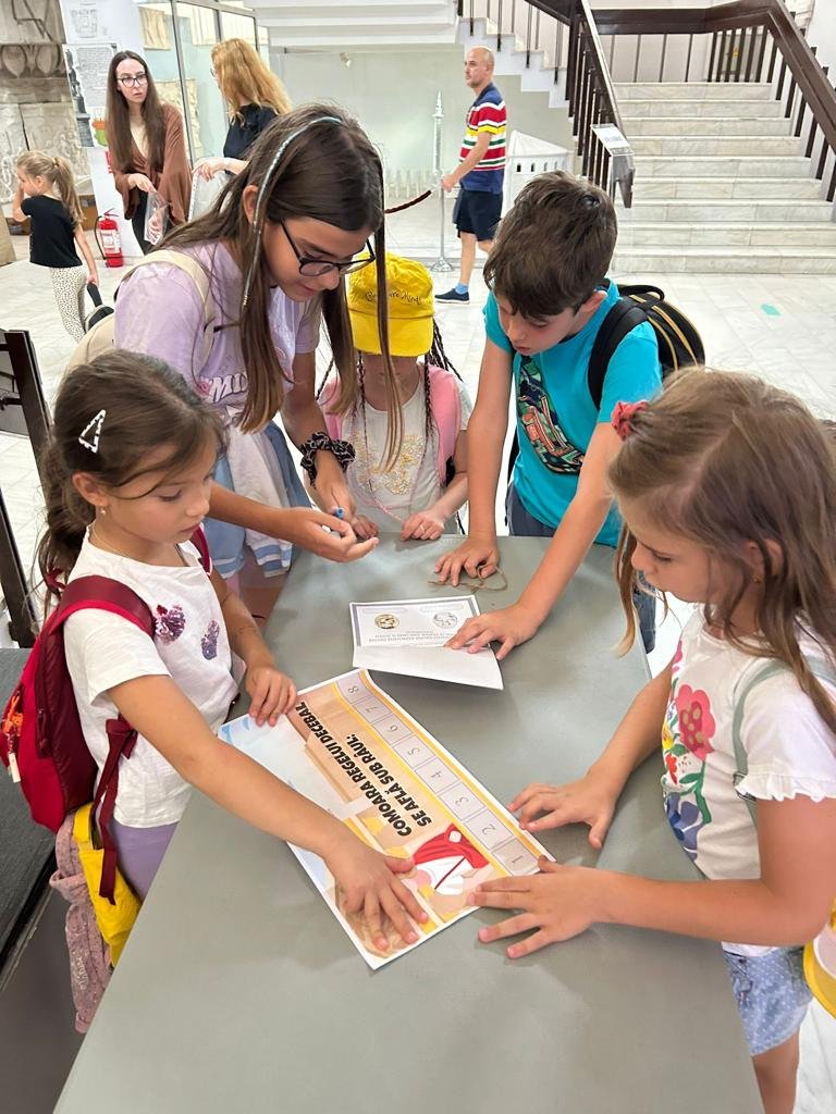 Group of children gathered around a table in a museum, engaged in an educational activity, using worksheets and papers. Background shows museum displays and stairs.
