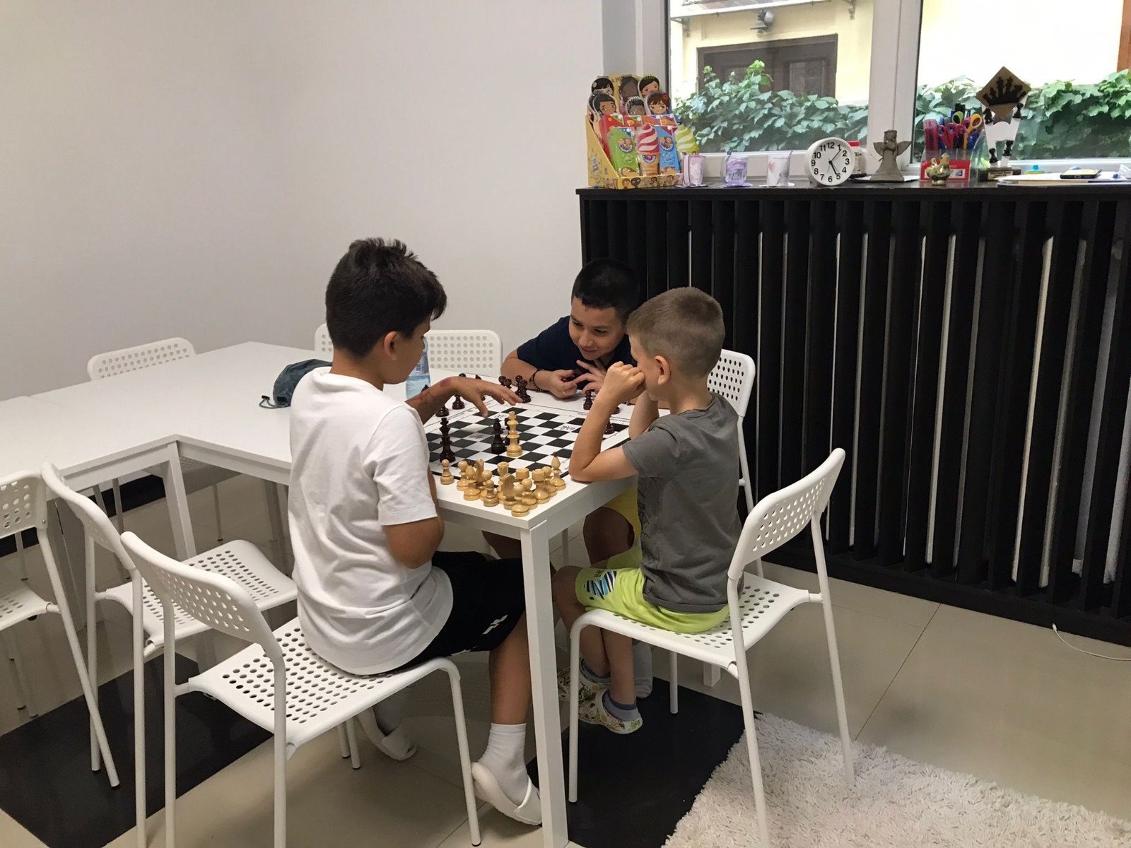 Three children playing chess at a white table in a room with white chairs and a window sill displaying decorative items.