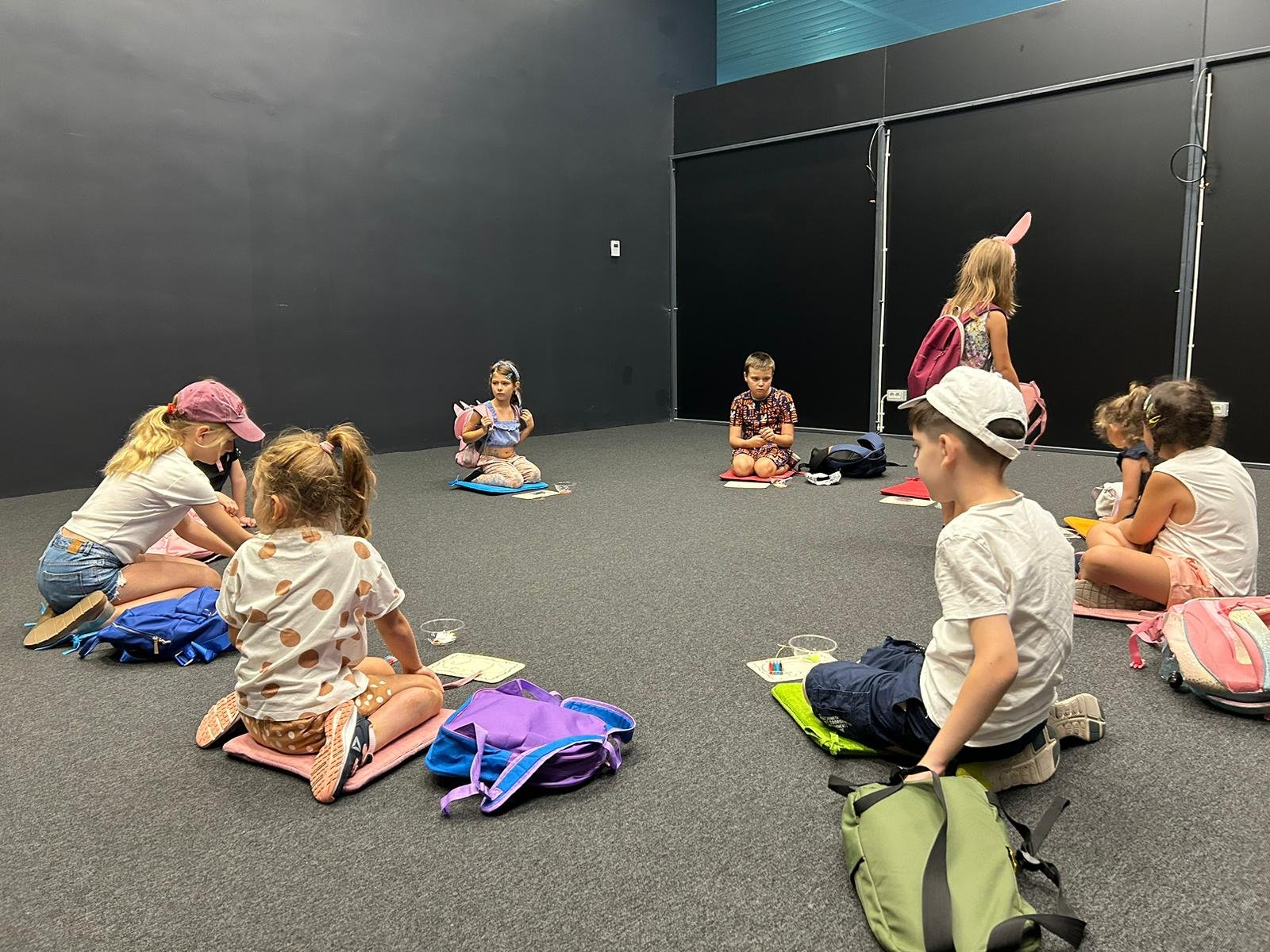 Children sitting on the floor in a classroom environment, with backpacks and notebooks around them.