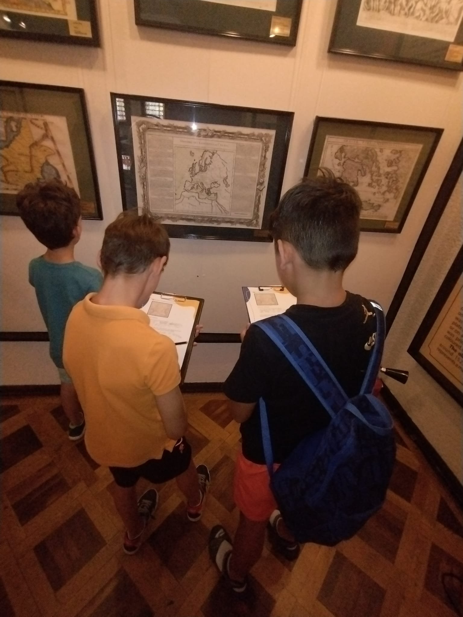 Three kids examining framed maps on a museum wall while holding clipboards.