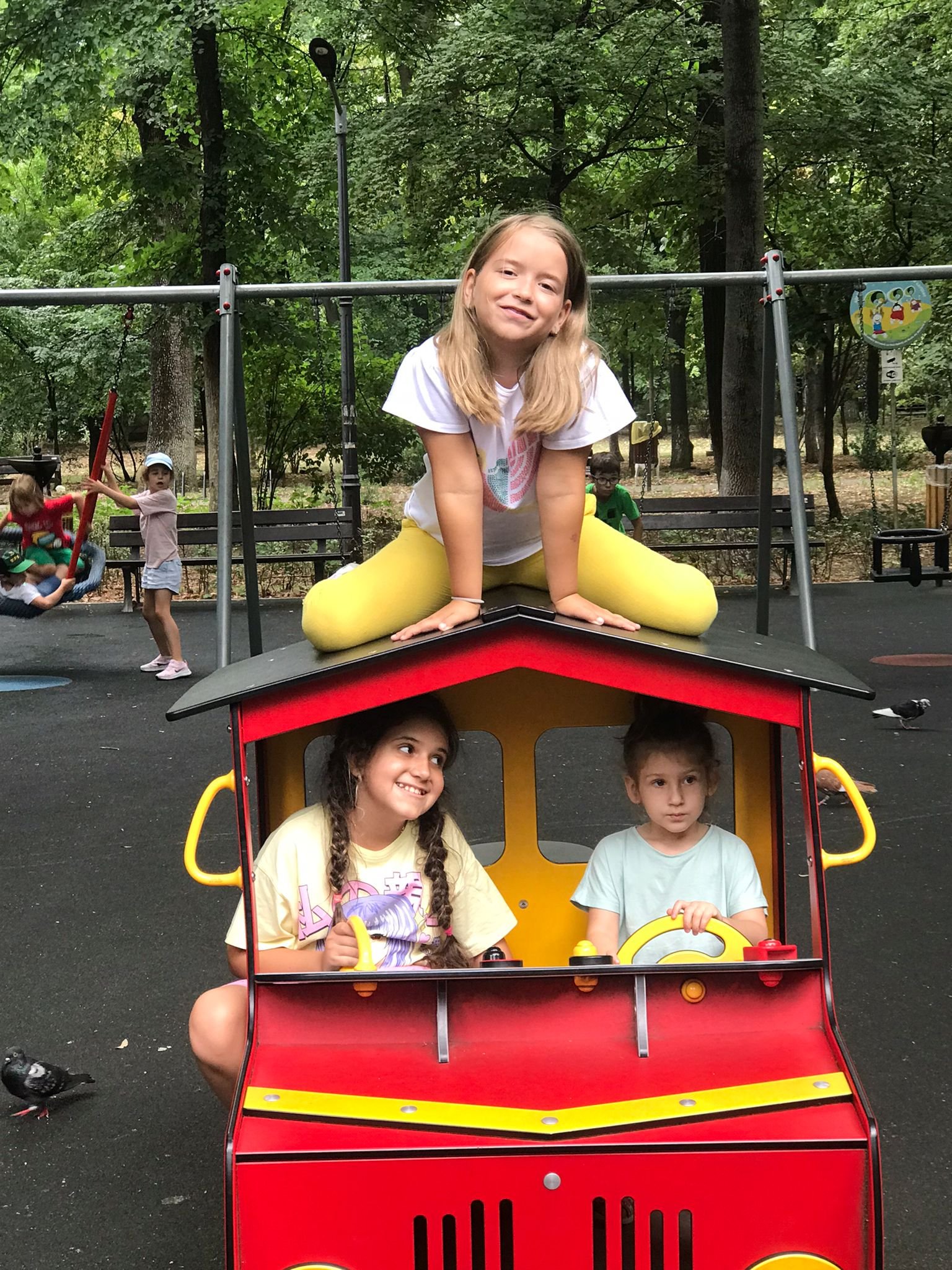 Children playing on a red and yellow playground toy resembling a vehicle, with one child sitting on top and two children inside. Trees and playground equipment are in the background.