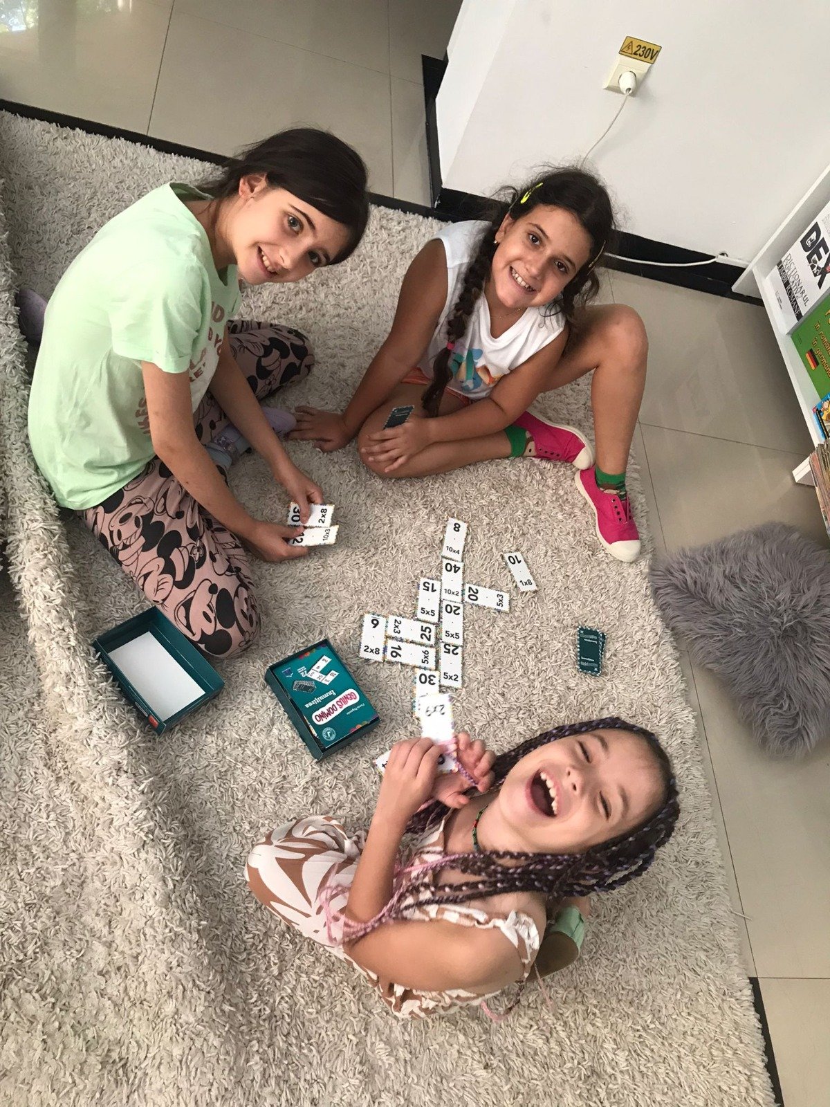 Three children sitting on a carpeted floor playing a math-based board game with cards and boxes.