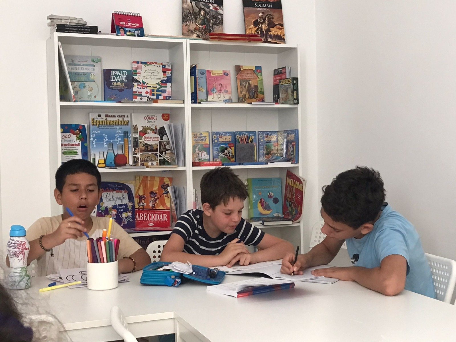 Three children sitting at a table engaged in drawing and writing activities, surrounded by colorful books on a white bookshelf in a classroom setting.