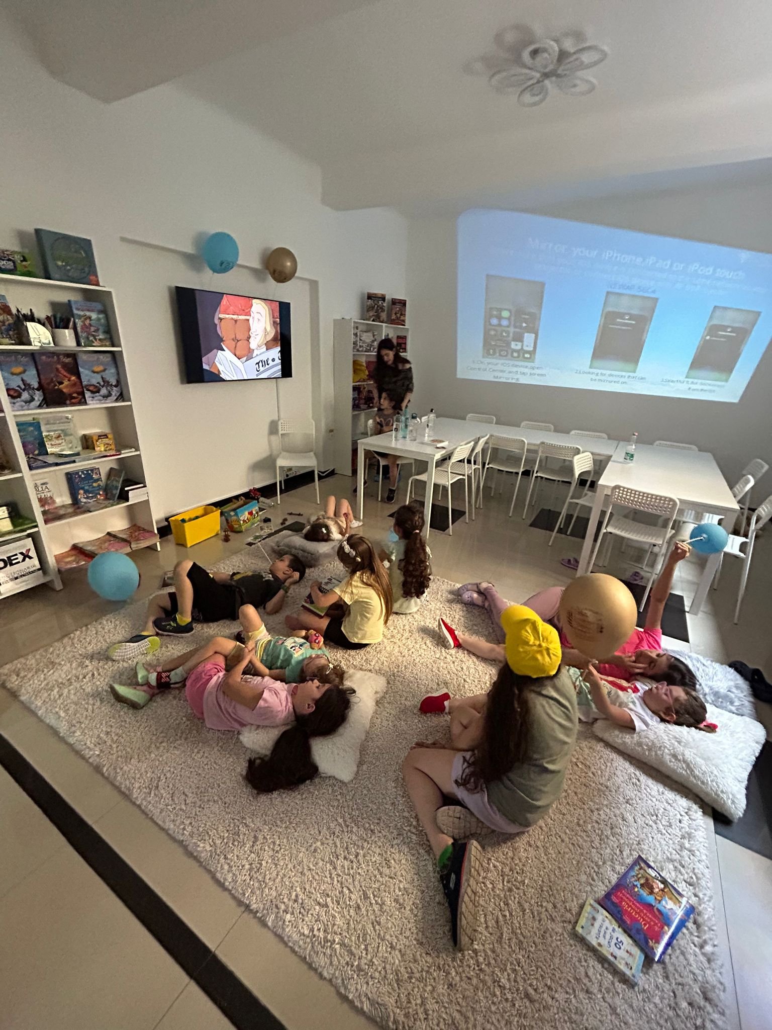 Children resting on a carpet while watching a movie in a room with a projector and bookshelves.