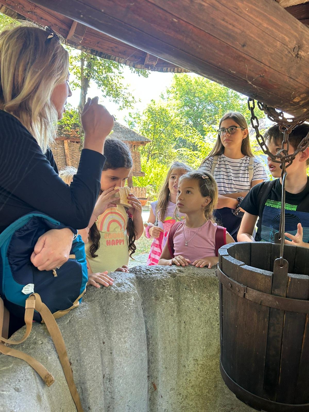 A group of children and an adult gathered around an old stone well, with a wooden bucket hanging by chains. The setting is outdoors, surrounded by trees and a wooden structure. One child is drinking from a cup, and others appear curious and engaged.