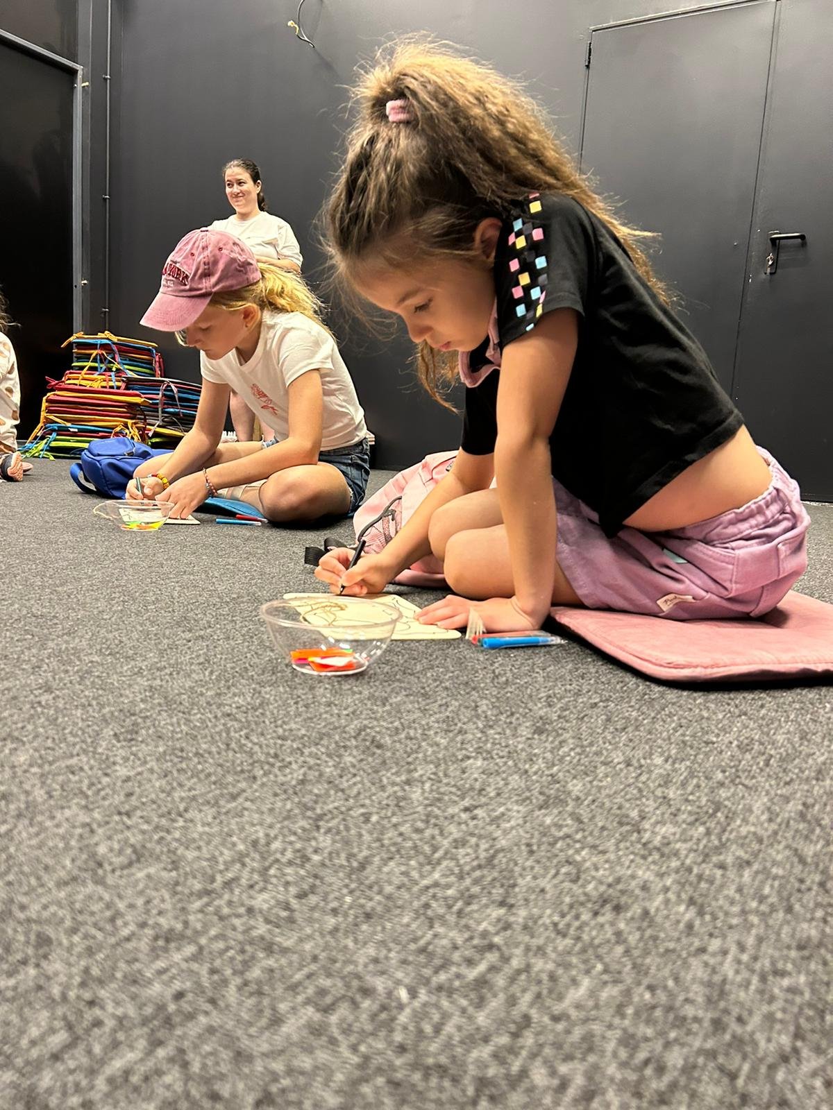 Children sitting on the floor engaged in art activity, coloring with markers, in a room with a dark wall and stacked chairs. An adult stands in the background.
